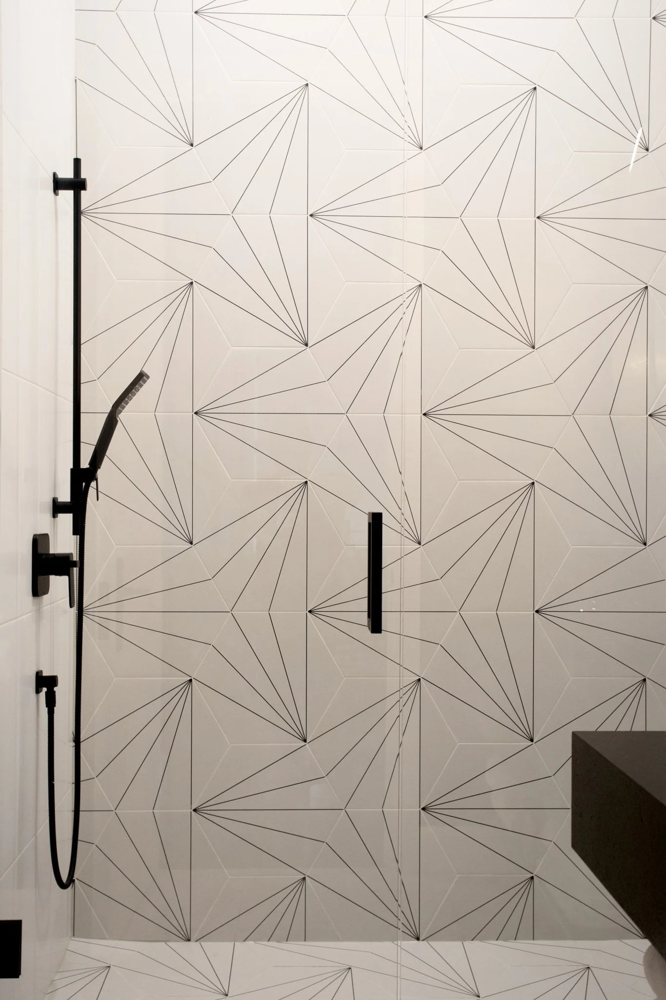 A modern shower area with a wall featuring geometric black line patterns on white tiles, a mounted black showerhead with a hose, and a black handle on the door.