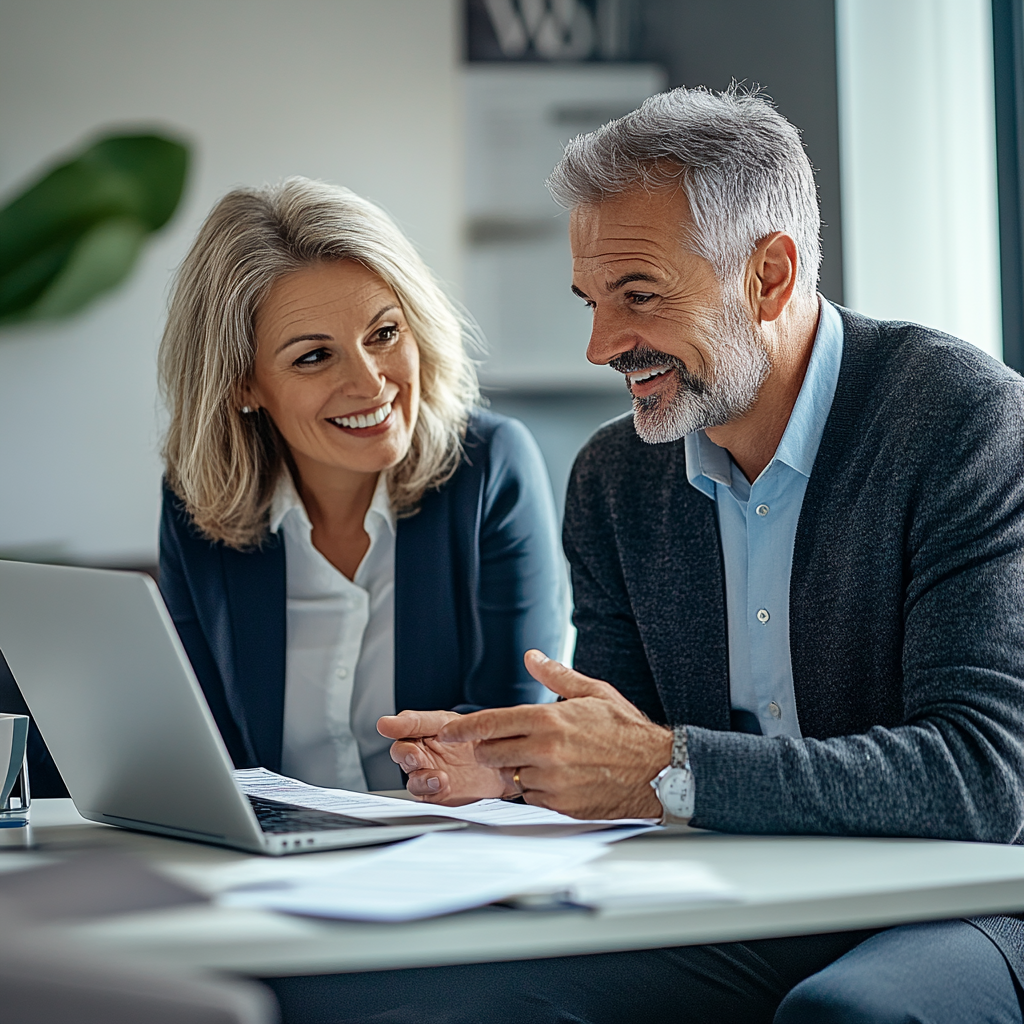 A man and a woman are sitting at a desk, smiling and having a conversation while looking at each other. The man gestures with his hands, and there is a laptop in front of them.