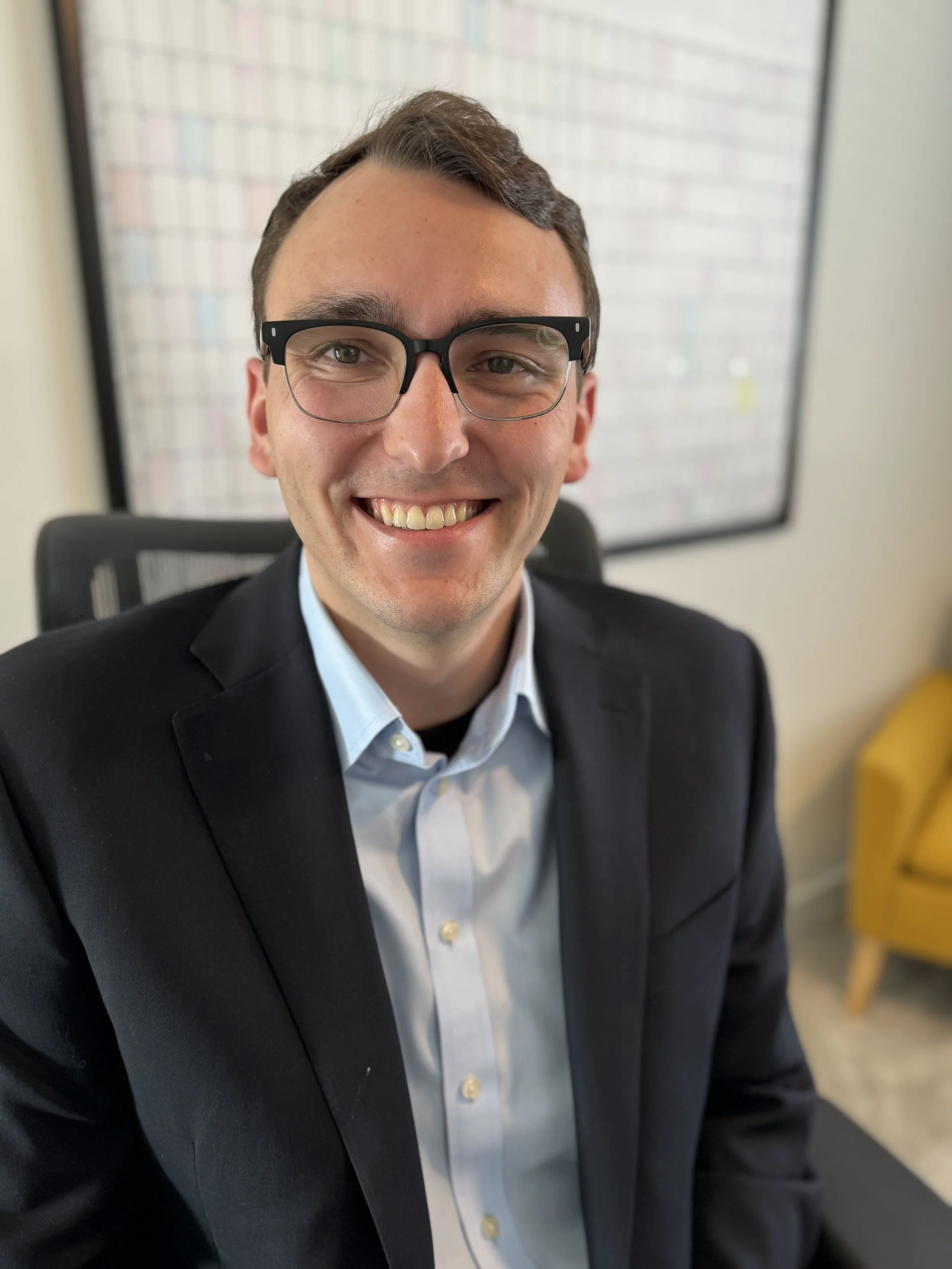 A smiling man in a dark suit and glasses sitting in an office chair, with a whiteboard behind him.