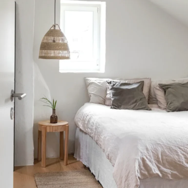 Einrichtung eines Schlafzimmers im Skandi-Stil mit weißem Bett, grauen Kissen, einem kleinen Nachttisch mit Pflanze, einer Wandlampe und einem Fenster im Dachgeschoss.