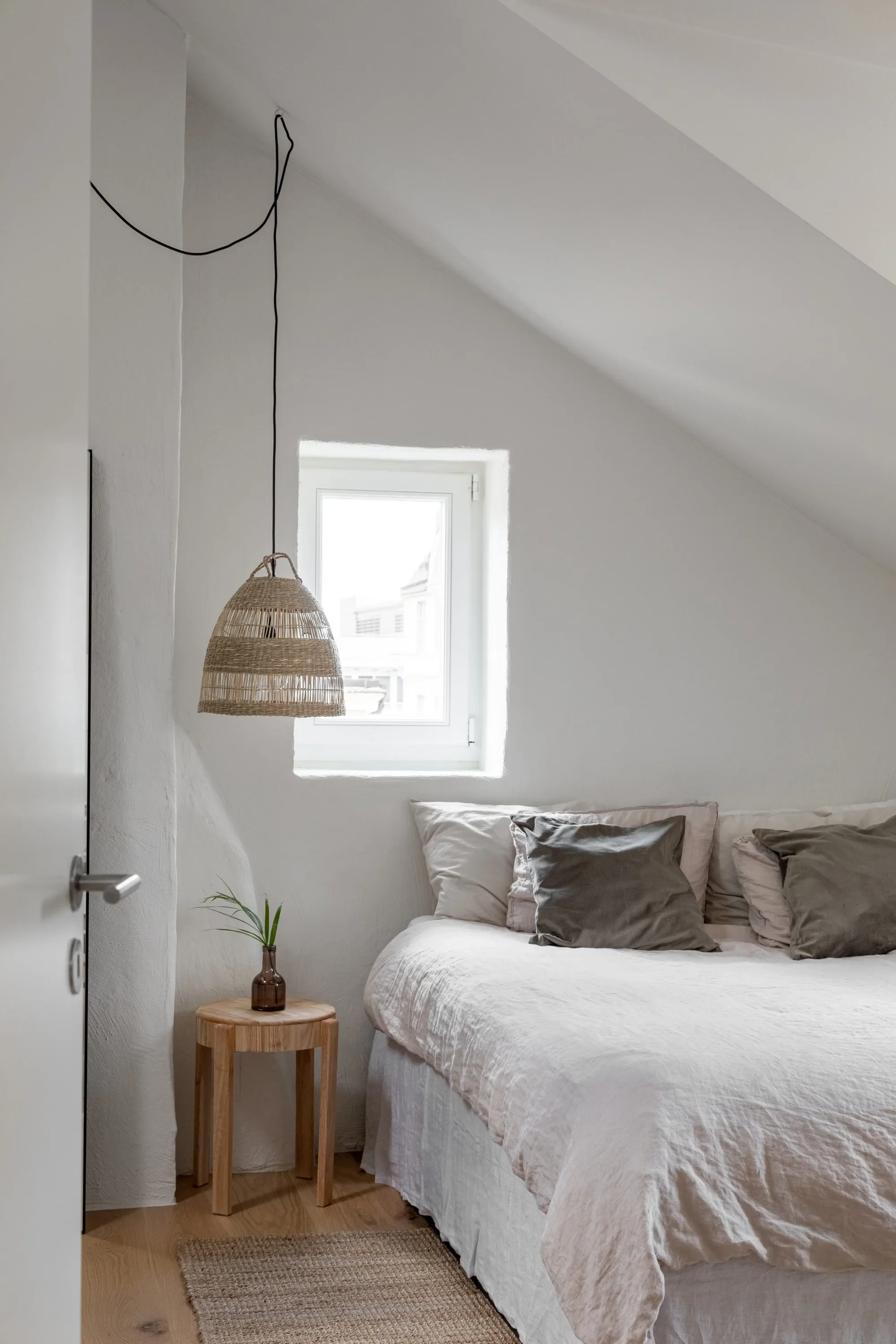 Schlafzimmer mit weißen Wänden, einem Bett mit weißen Bettdecken und grauen Kissen, einem natürlichen Holznachtisch mit einer Vase und einer kleinen Pflanze, einer hölzernen Hängelampe, einem kleinen Fenster, einem Teppich auf dem Holzboden.