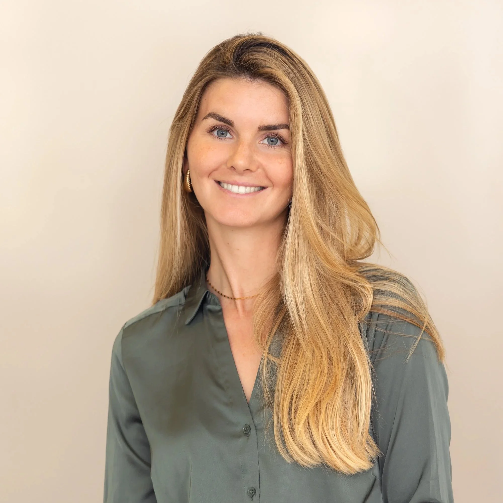 Eine junge Frau mit langem, blondem Haar, blauen Augen, und einem freundlichen Lächeln, trägt eine graue Bluse und goldene Ohrringe, steht vor einem neutralen Hintergrund. Innenarchitektin Jule Huschka. Interior Designerin bei Onterior Design.
