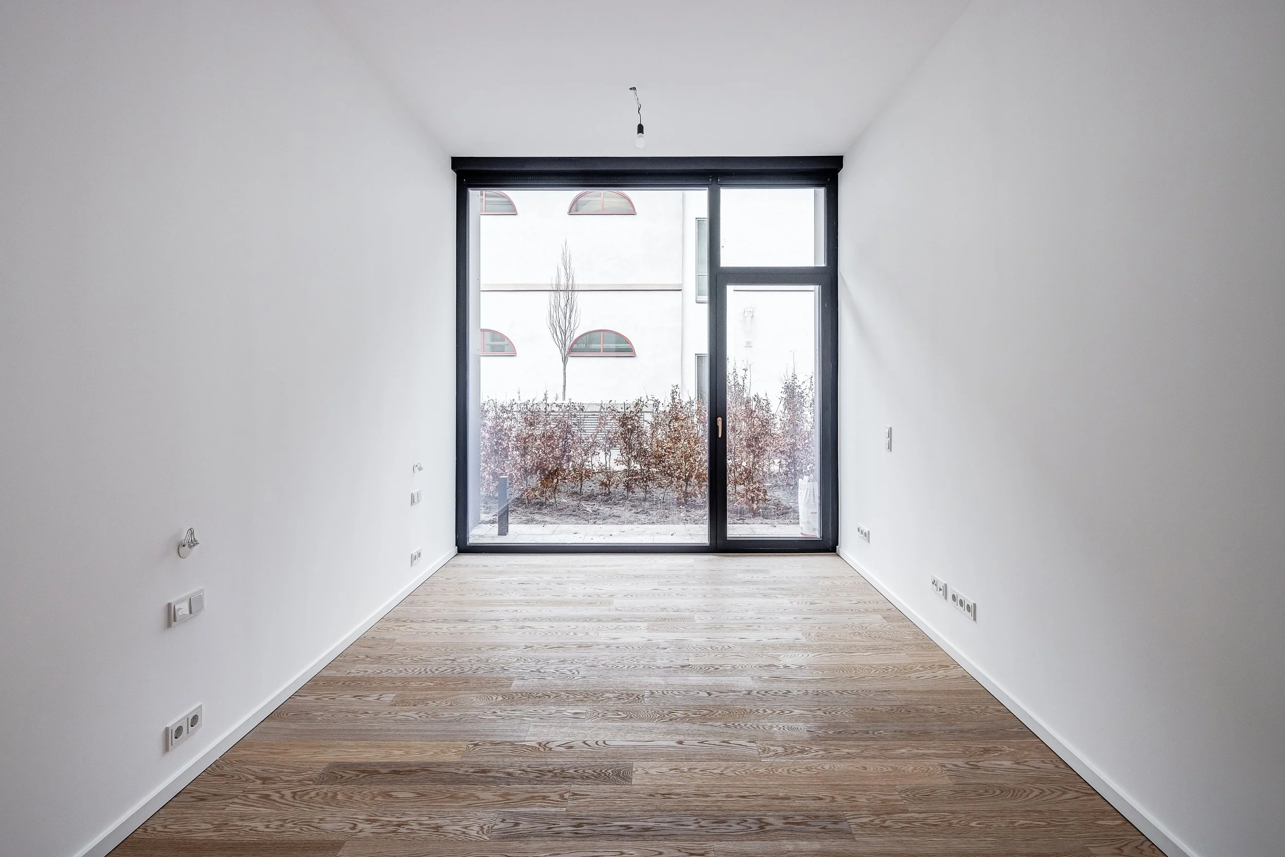 Leeres, modernes Zimmer mit weißen Wänden, Holzboden und großen Fenstern nach draußen. Einrichtungsberatung fuer 1-Zimmer-Apartment in München. Online Designberatung. Innenarchitektur und Interior Design Beratung und Planung mit Onterior. 