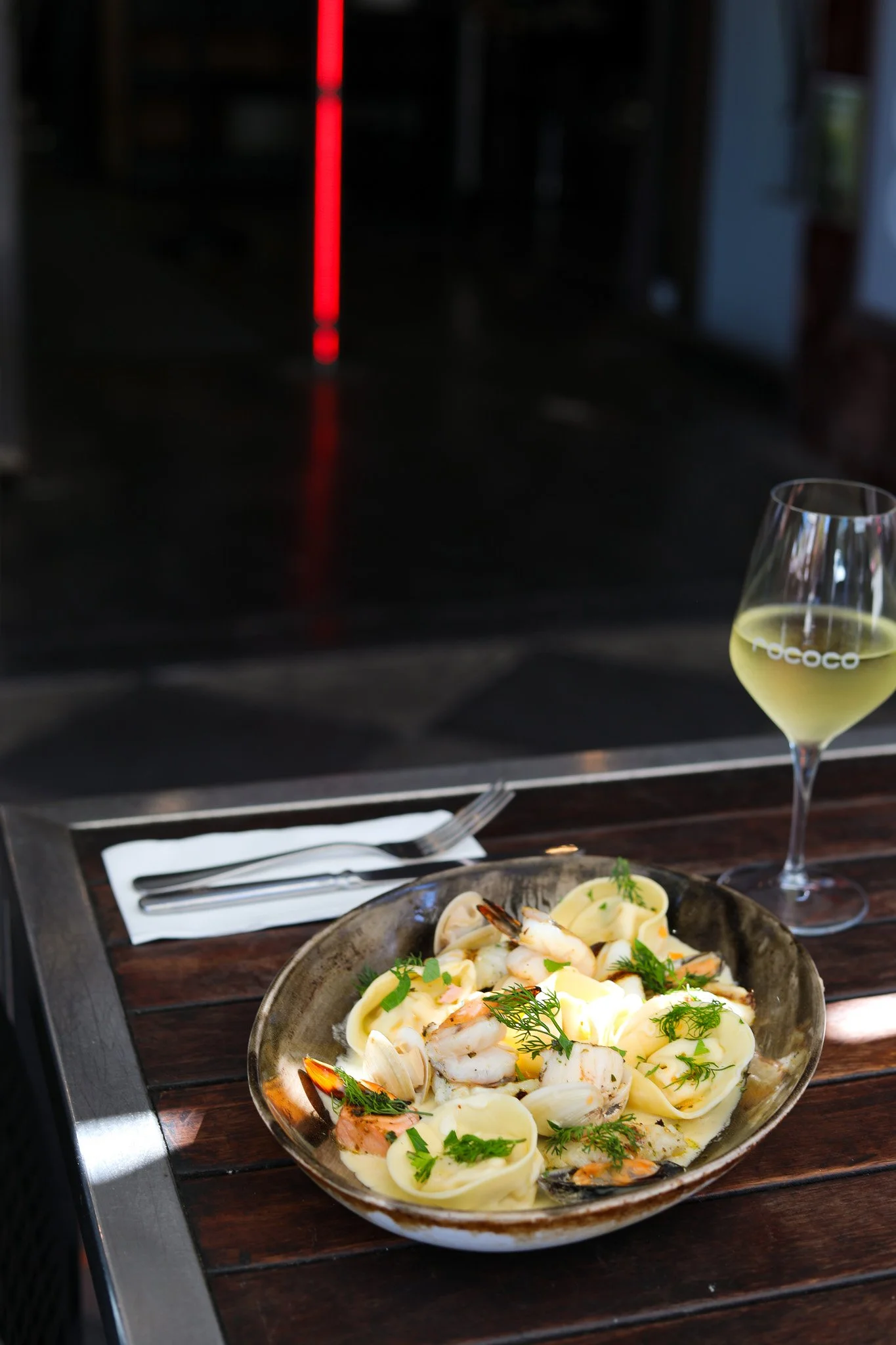A bowl of seafood pasta with shrimp, clams, and herbs on a wooden table, with a glass of white wine and silverware nearby, in a restaurant setting.