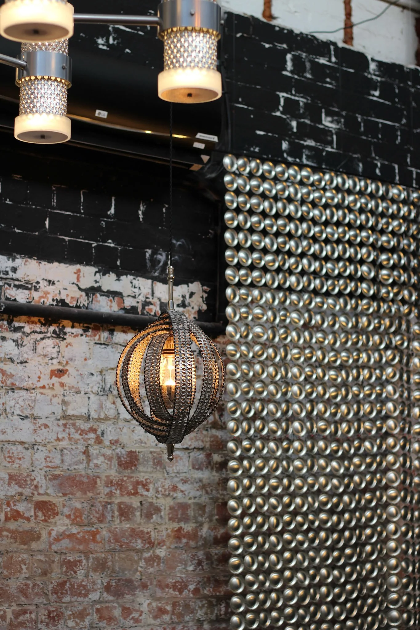 Decorative lighting fixtures hang in front of a brick wall painted black and white, with a grid of metallic balls on the right.