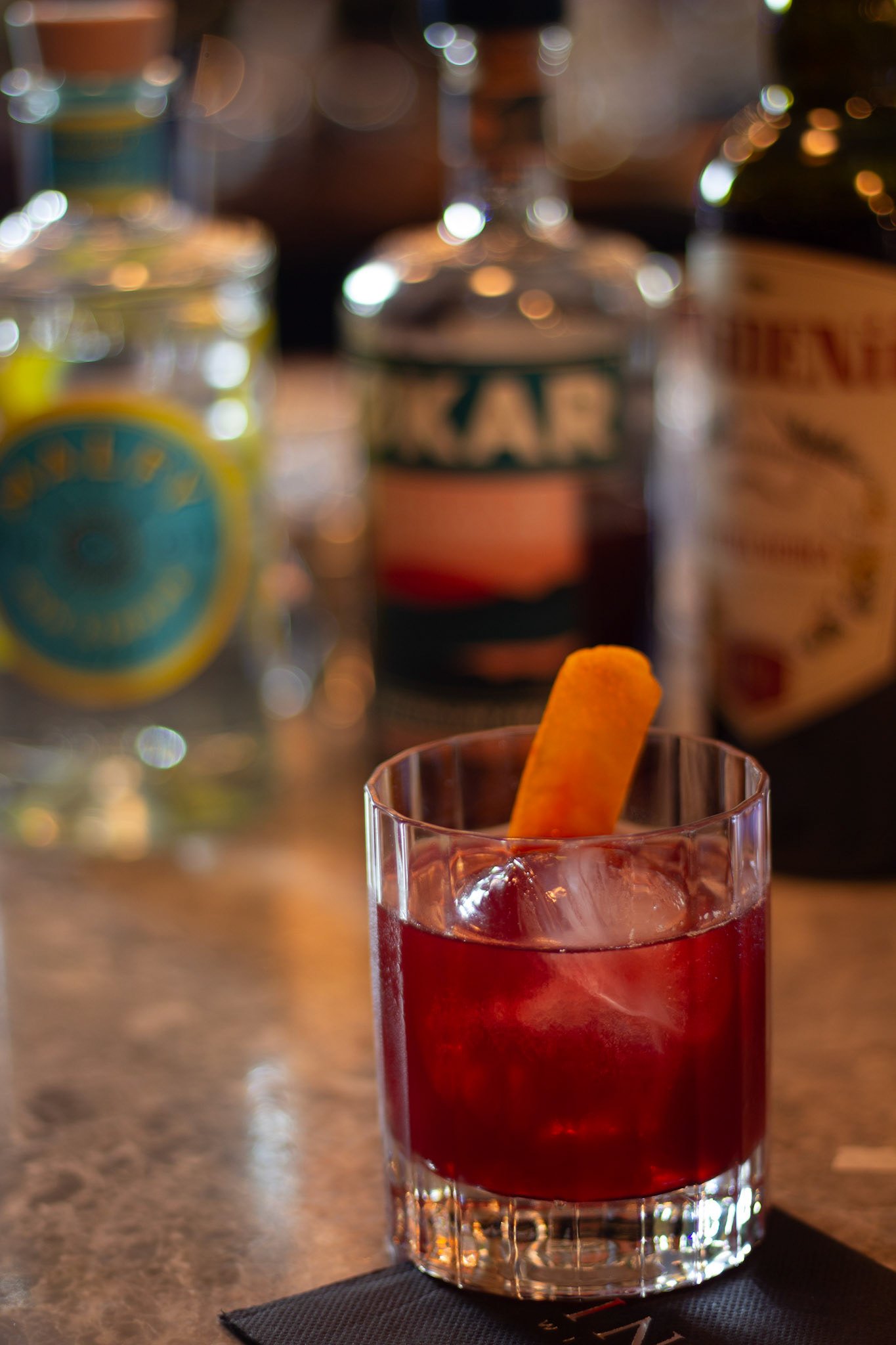 A close-up of a red cocktail in a glass with a yellow fruit garnish and ice, on a black napkin on a wooden bar counter with blurred bottles and glasses in the background.