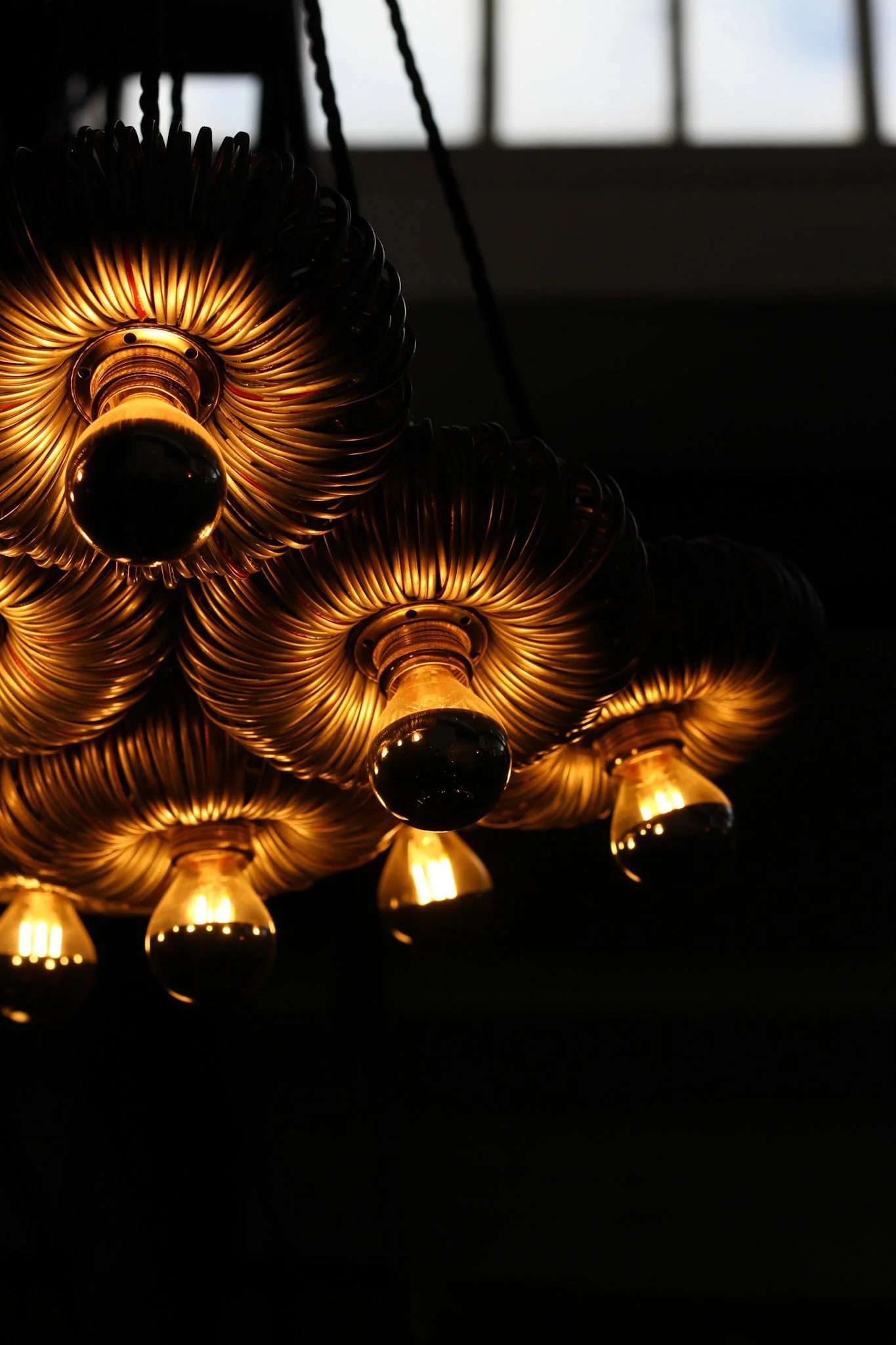 Close-up of a modern chandelier with gold spiral design and round black accents, illuminated against a dark background with a window in the distance.