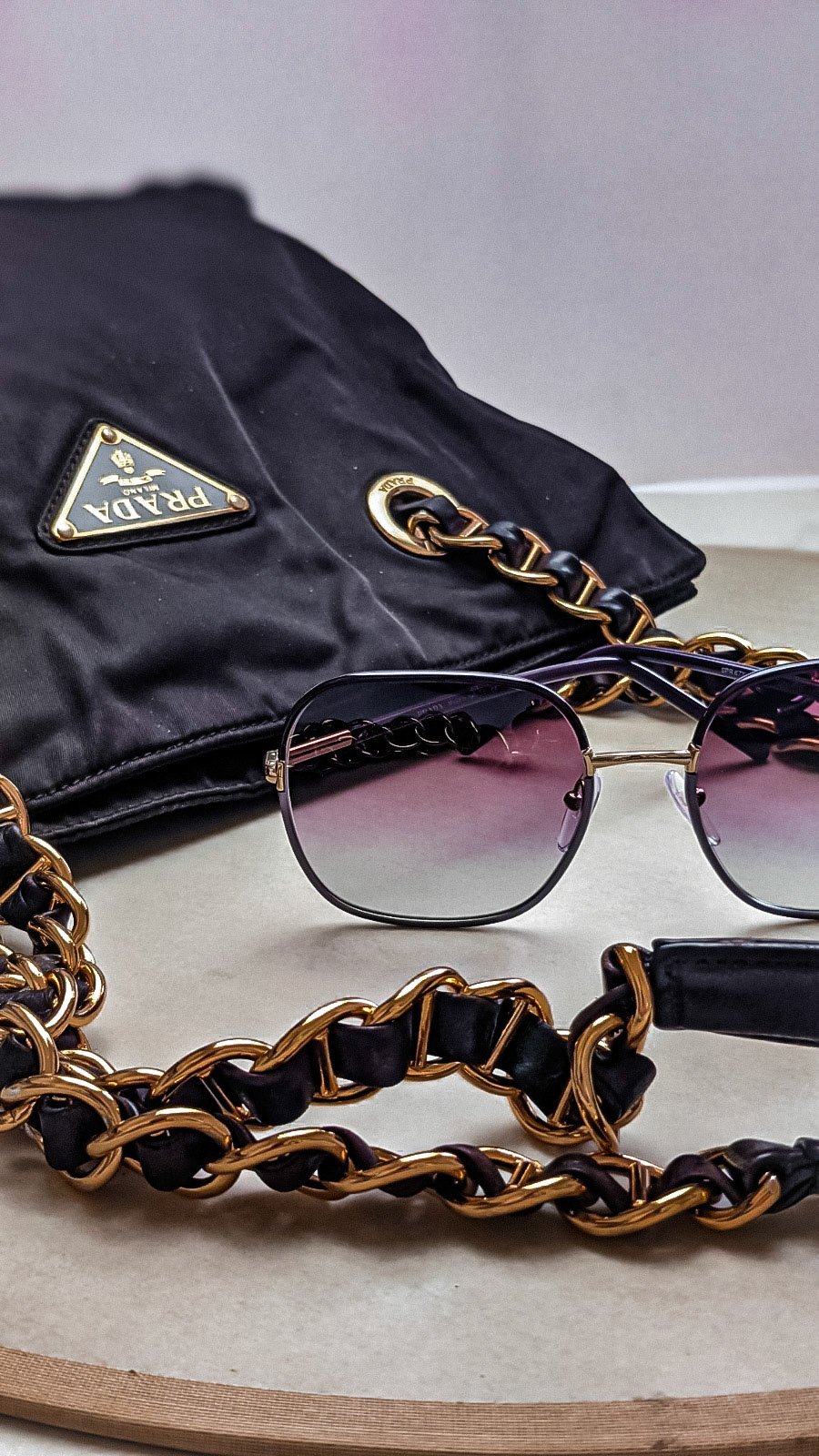 Black Prada handbag with a gold chain strap and logo, pair of sunglasses with gradient lenses, and a gold and black chain accessory, all placed on a beige surface.