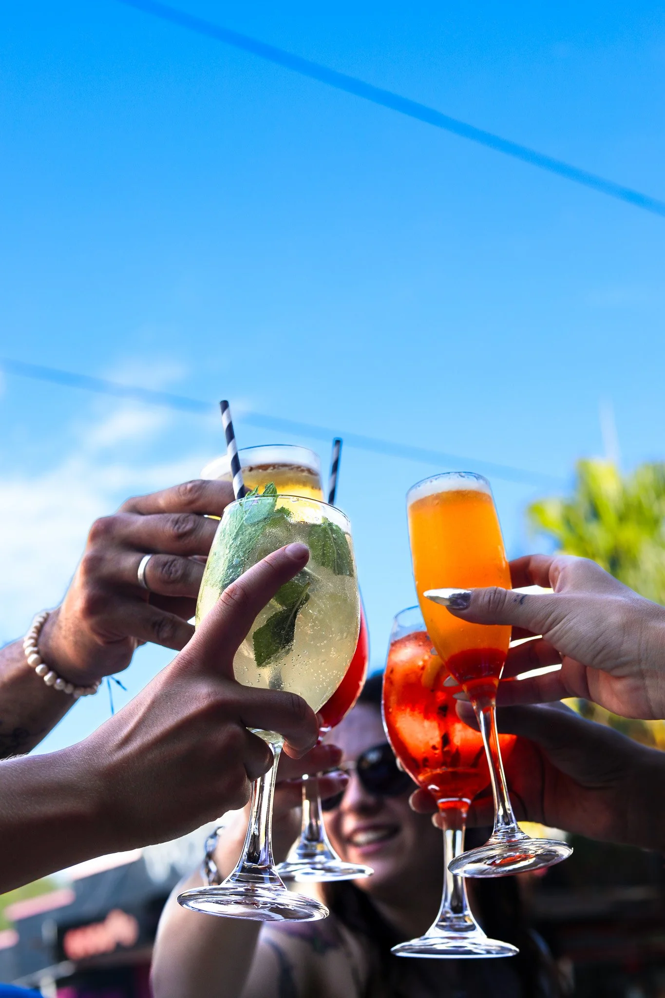Multiple people holding up colorful cocktails in a toast against a blue sky.