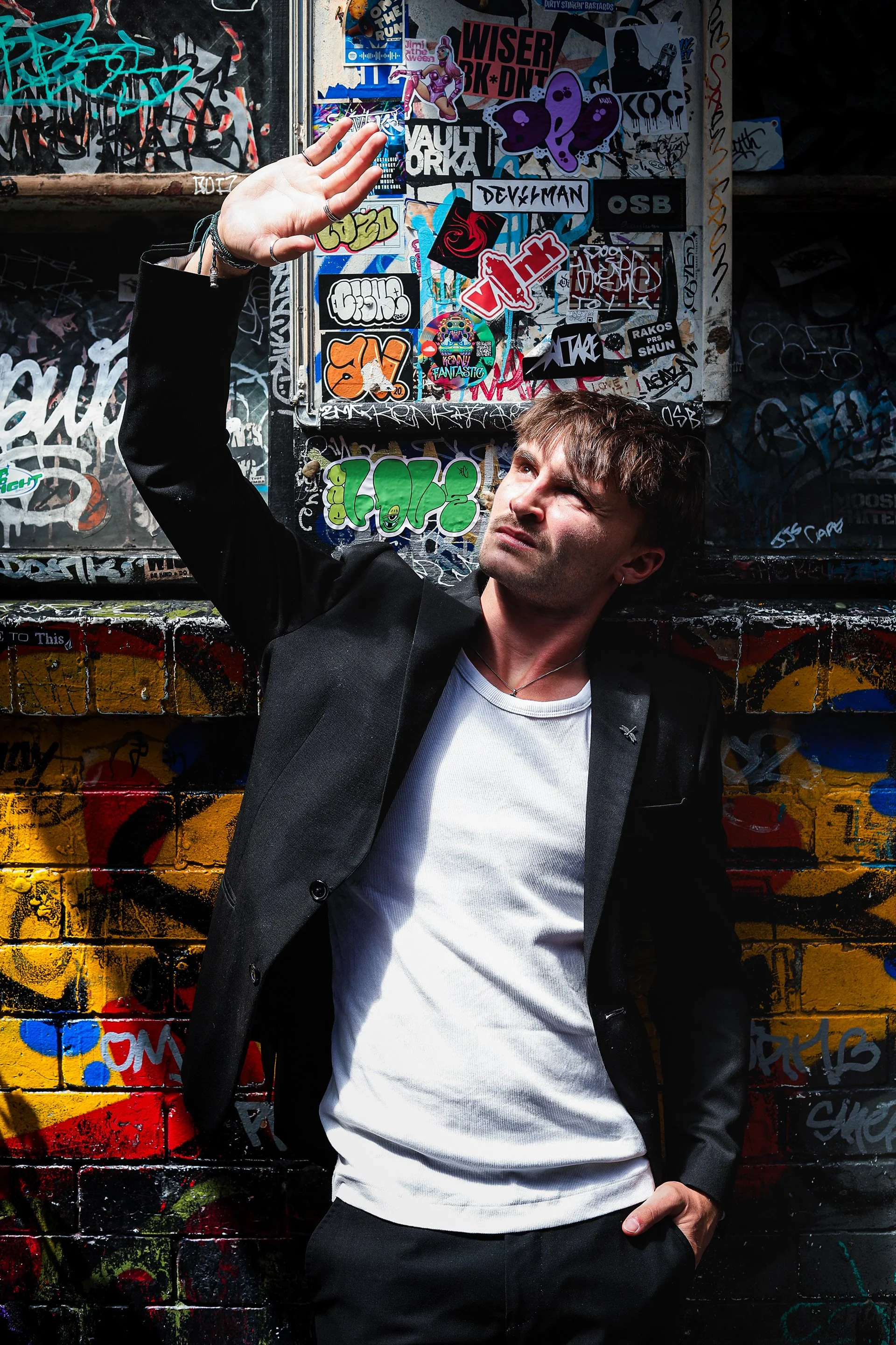 A young man with brown hair wearing a black blazer and white shirt is standing against a colorful graffiti-covered wall, holding his hand up as if shielding his eyes from the sun while looking upwards.