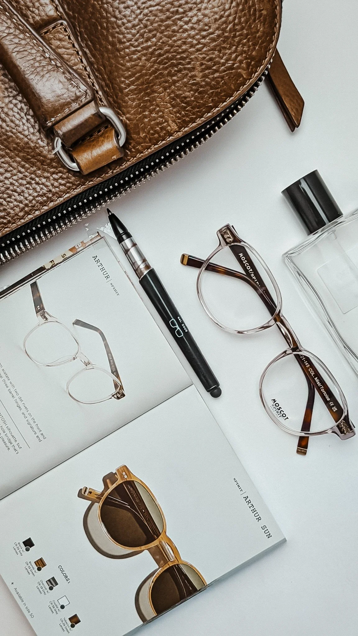 A close-up of a leather backpack in brown and tan, with a zipper, a black pen, a pair of eyeglasses with tortoise shell frames, a bottle of perfume, and a sunglasses catalog or magazine on a white surface.