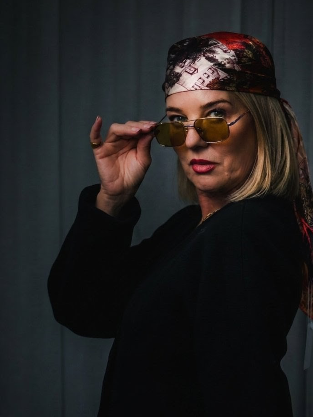 A woman wearing a colorful headscarf, black jacket, and stylish yellow-tinted sunglasses looks over her shoulder, adjusting her sunglasses with her right hand, against a dark background.