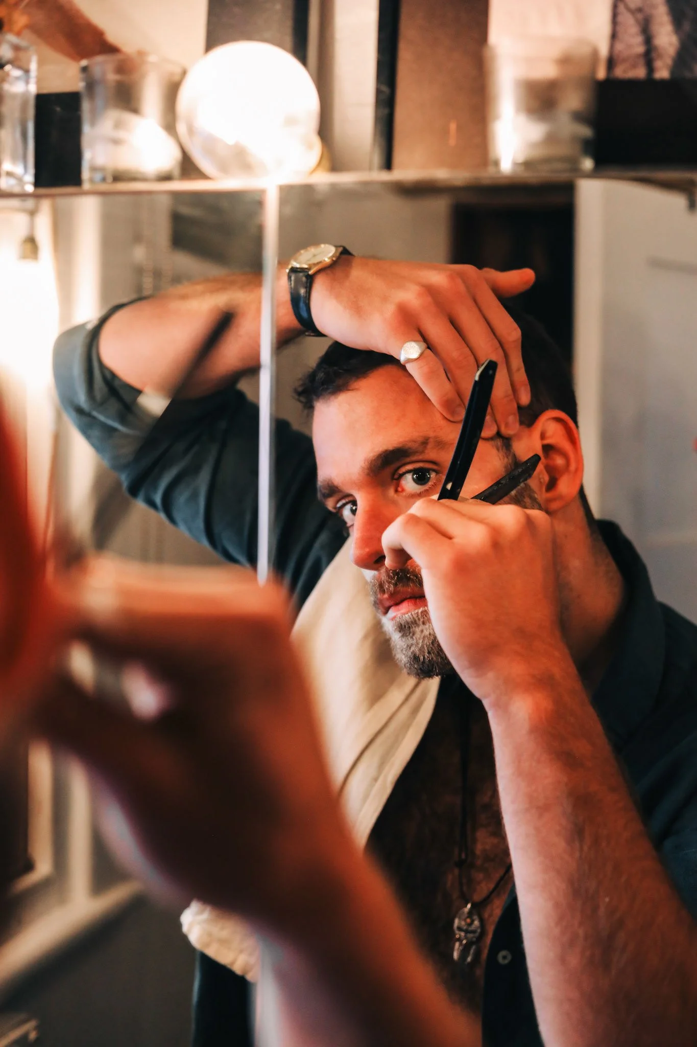 A man with a beard and mustache getting a haircut under a mirror, with a barber holding scissors and comb.