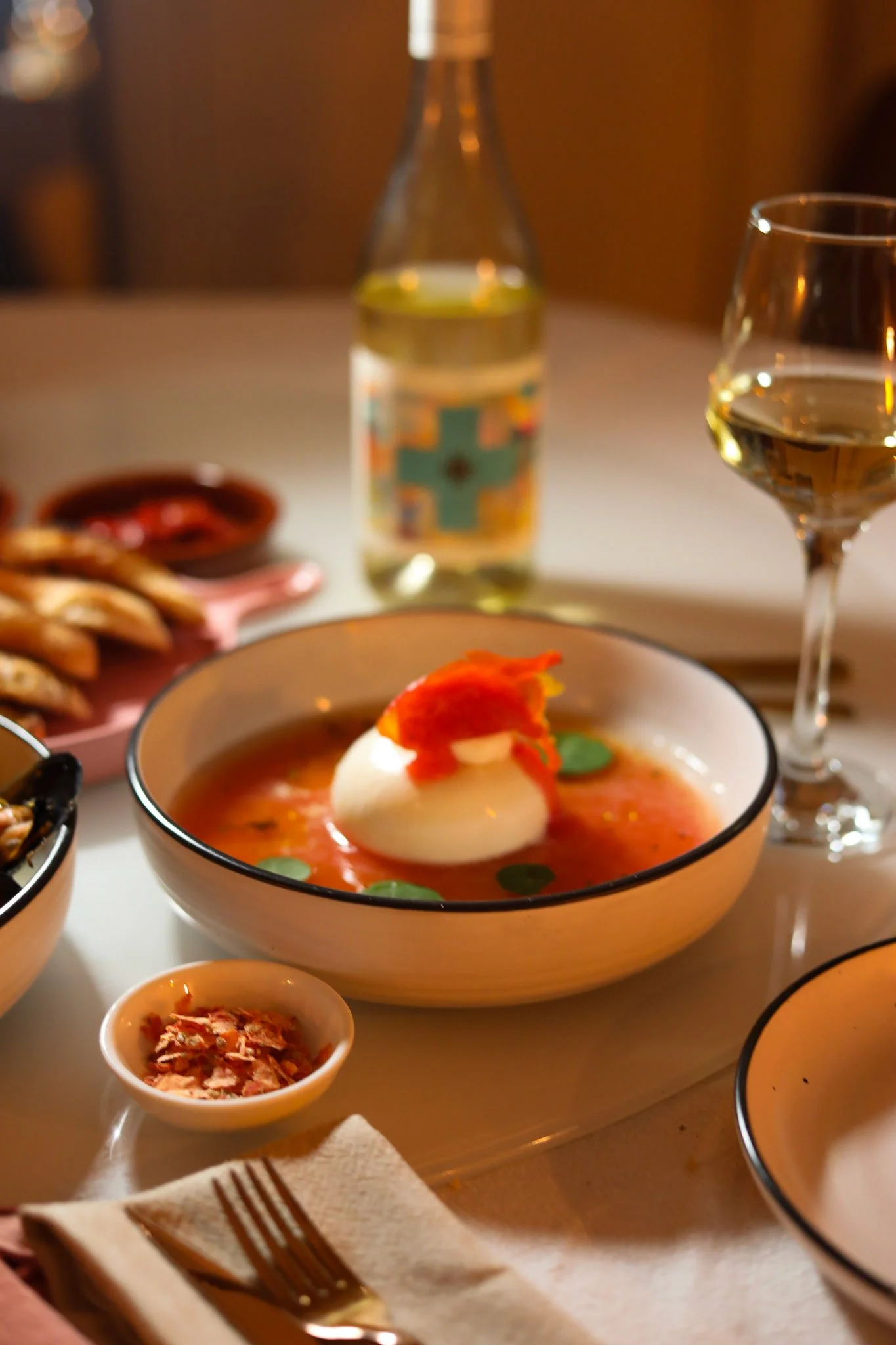 A table set with a bowl of poached eggs with red sauce and green herbs, a glass of white wine, a bottle of white wine, and small dishes of salsa or sauces.