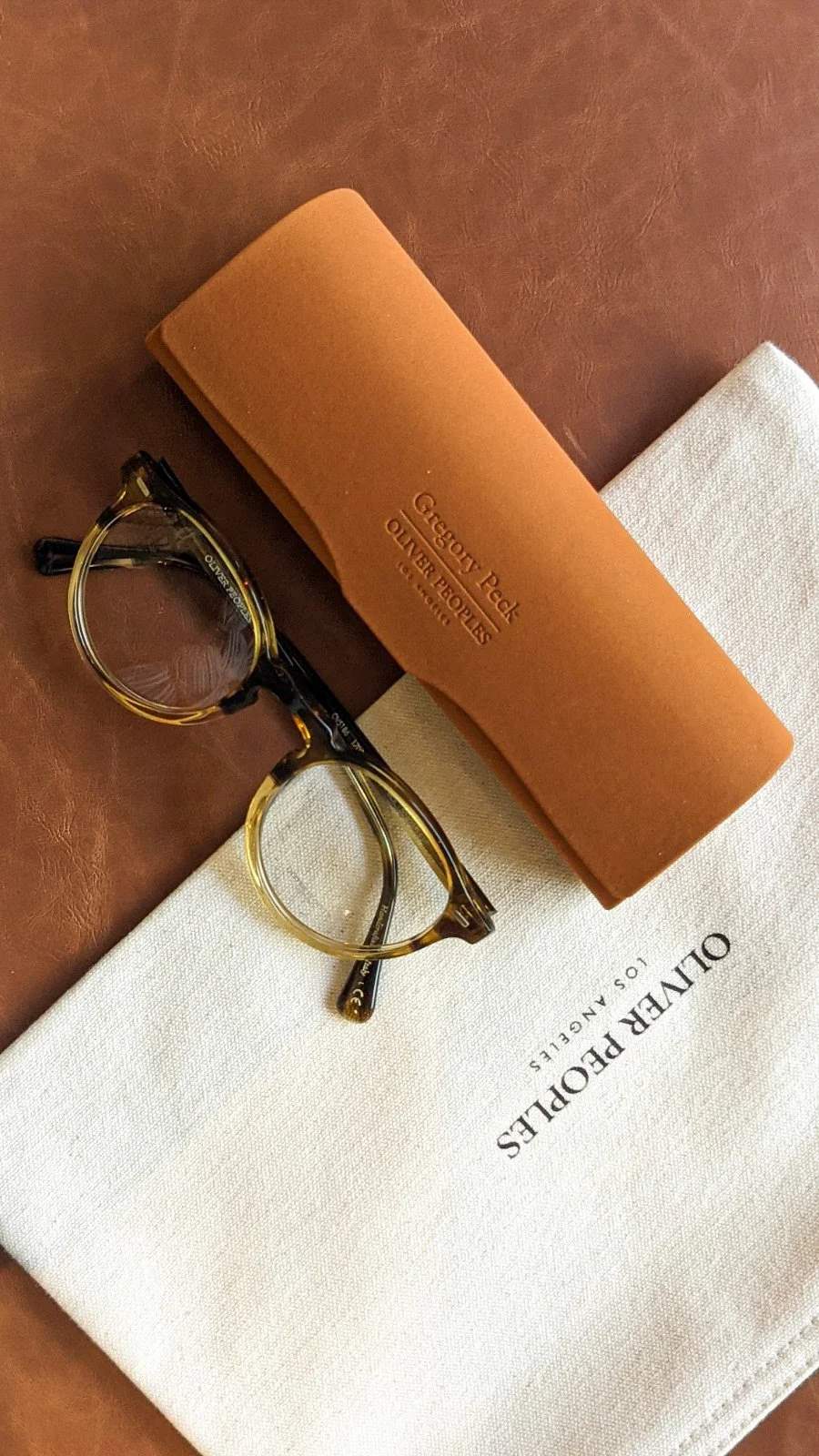 A pair of tortoiseshell reading glasses with a black and yellow frame, resting on a cream-colored cloth with "OILNER POLLIES" written on it, next to a tan eyeglass case with "GREGORY PECK OLIVER PEOPLES" embossed on it, all on a brown surface.