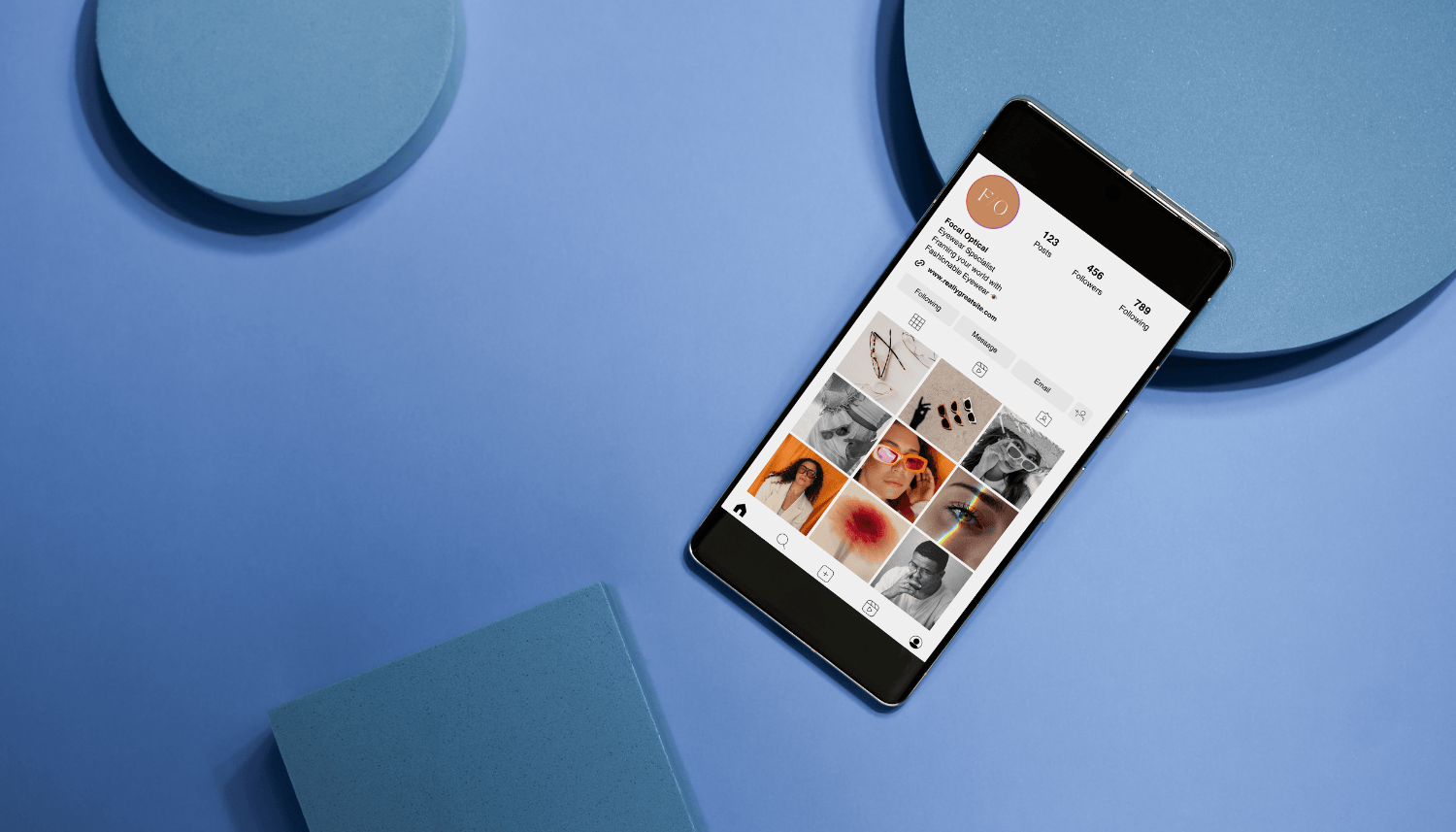 Smartphone displaying an Instagram profile with various photos, placed on a blue surface with blue decorative objects around it.