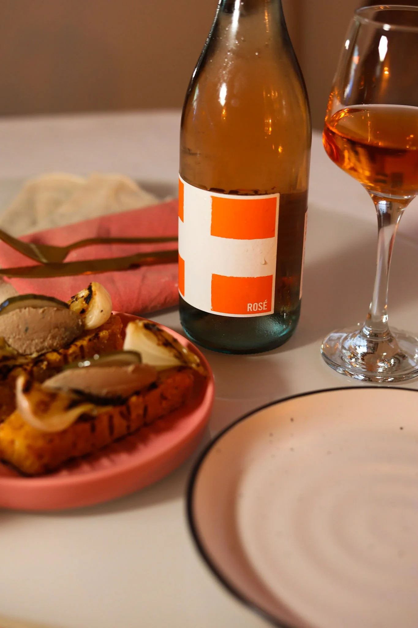 A table setting with a pink bowl of grilled eggplant slices topped with onions, a glass of rosé wine, a brown bottle of rosé wine, a pink napkin, and cutlery, with a white plate and a brown background.