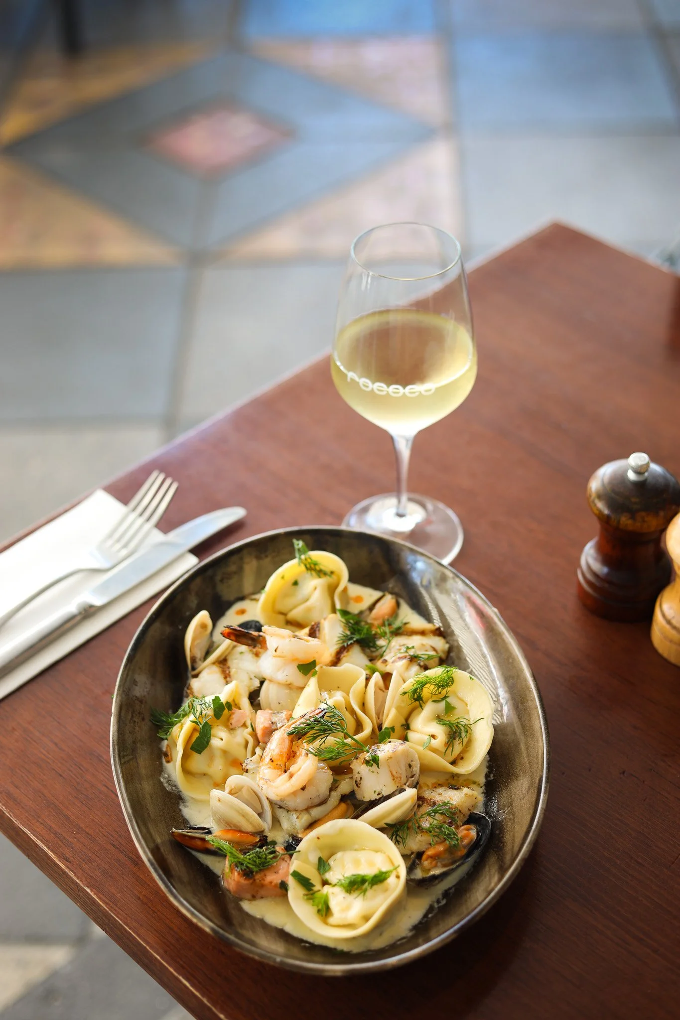 A plate of seafood pasta with shrimp, mussels, and garnished with herbs, next to a glass of white wine on a wooden table in a restaurant setting.