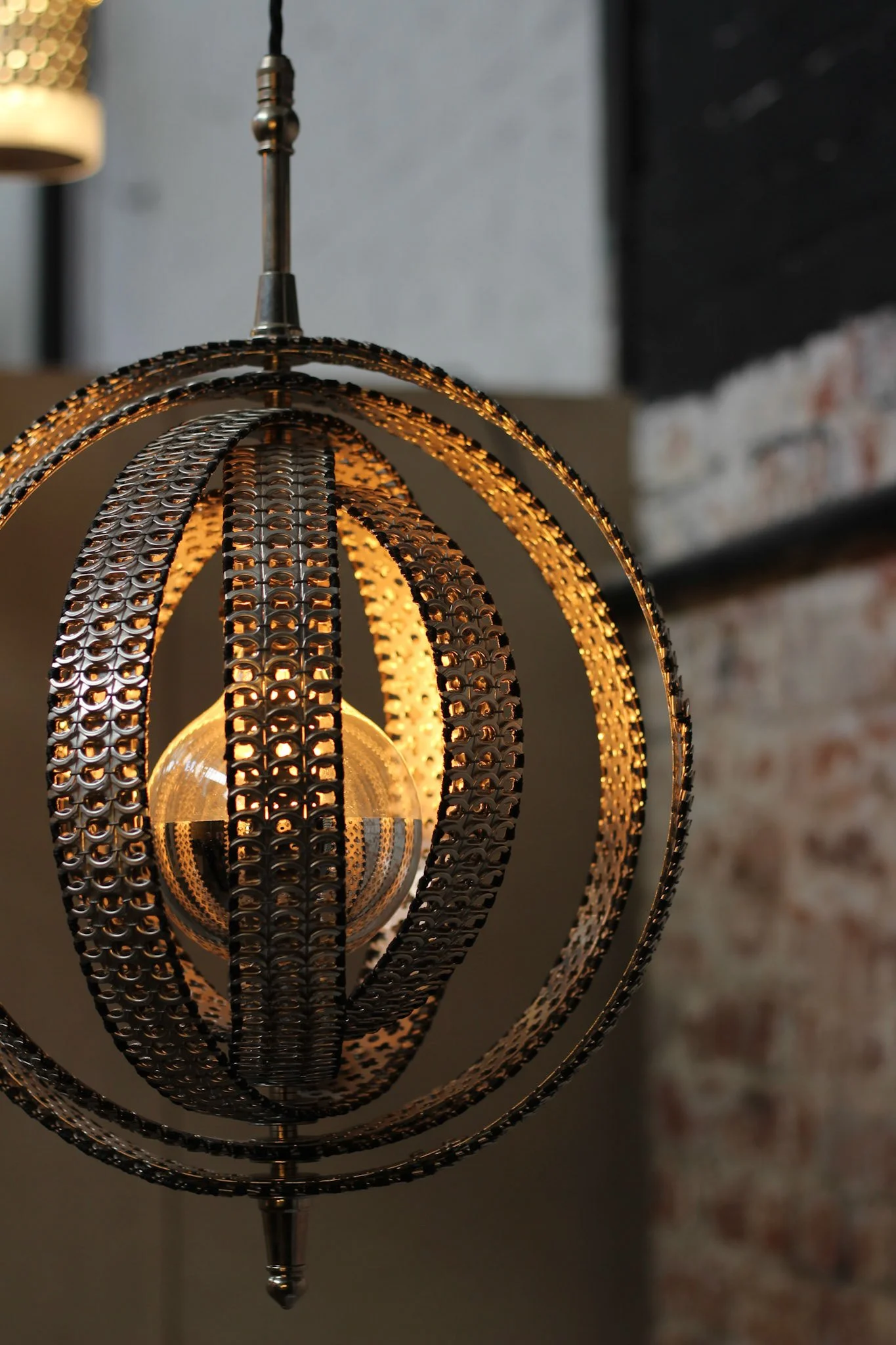 A close-up of a modern, metallic pendant light fixture with circular rings illuminated from within, hanging in a room with a brick wall background.