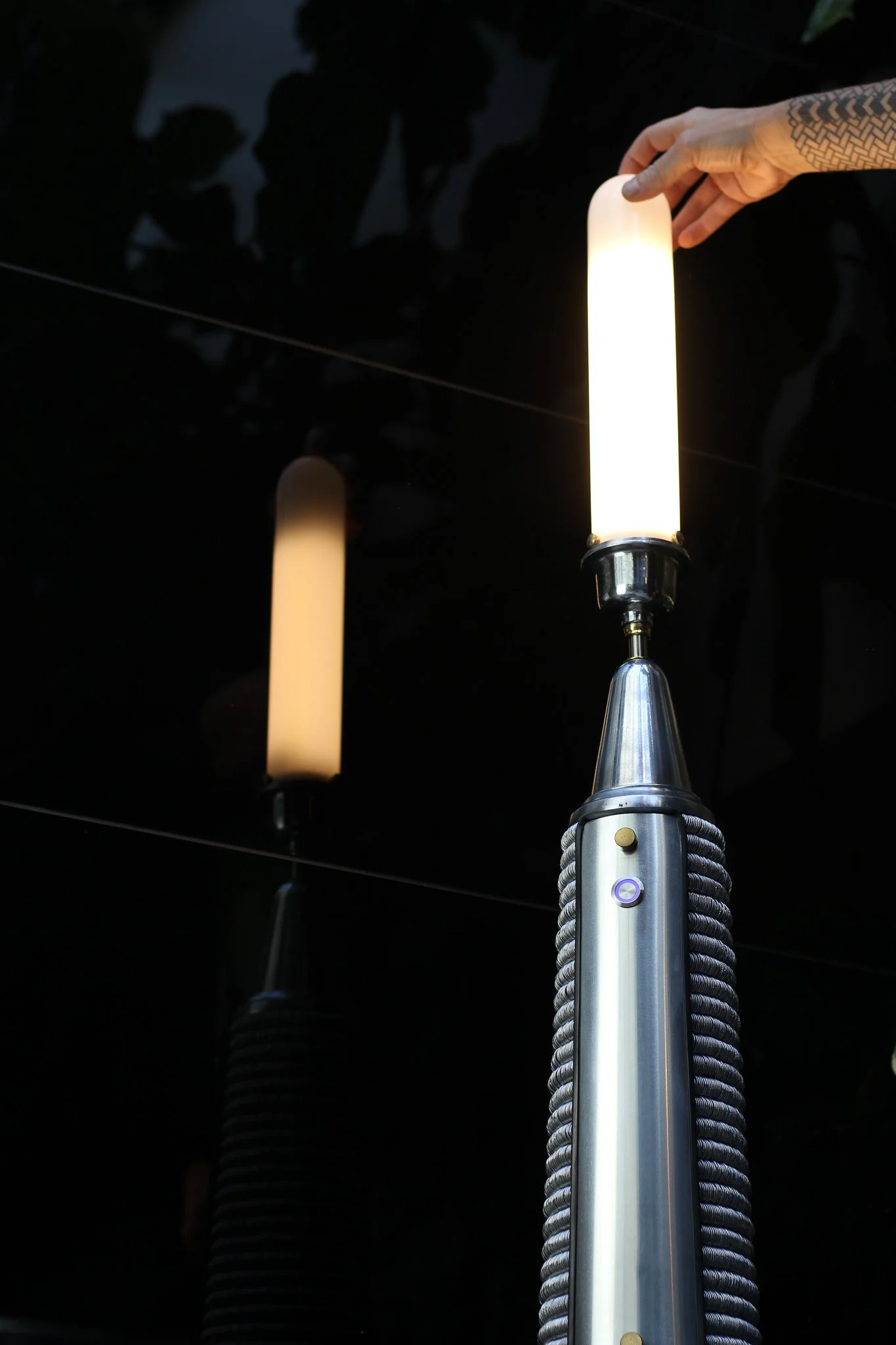 Close-up of a person's hand adjusting a cylindrical, glowing, white light fixture with metallic base, mounted on a metallic stand with coil springs. Reflection of the light fixture is visible on a dark, glossy surface.
