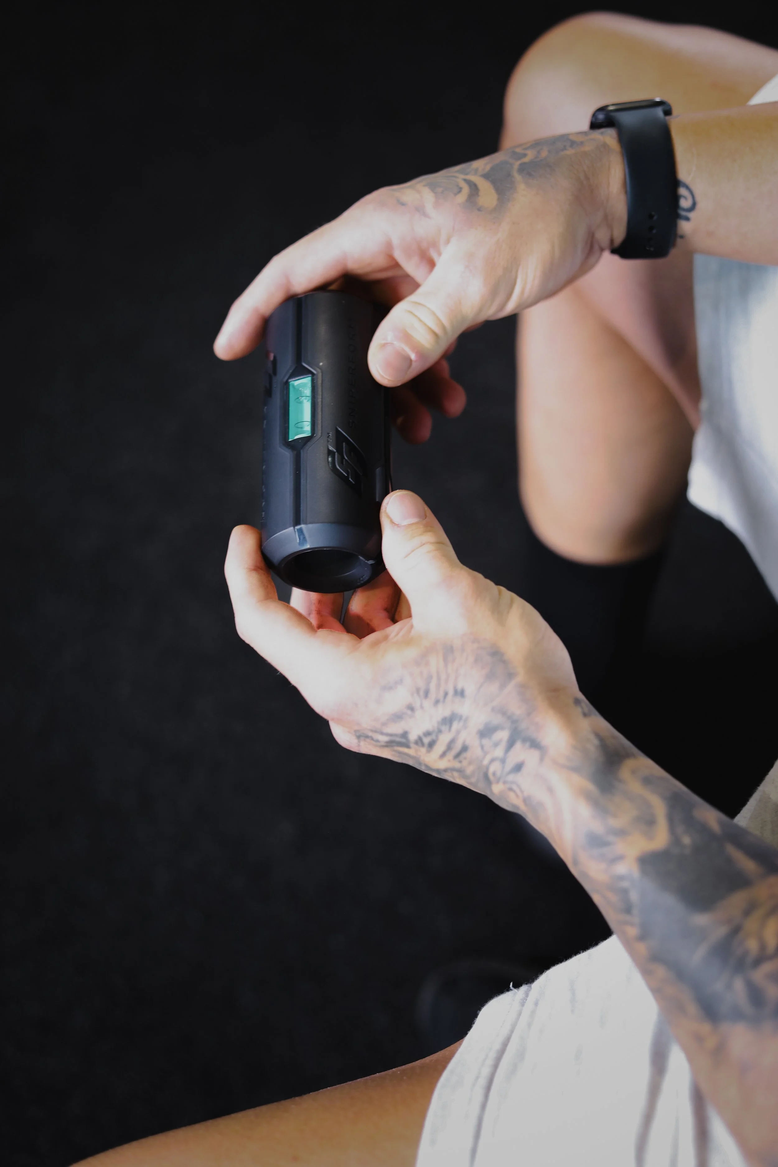A person with tattoos on their left hand and wrist, wearing a black fitness tracker, holding a black wireless fitness device while resting their right knee on a black surface. The person is wearing a gray top and white shorts.