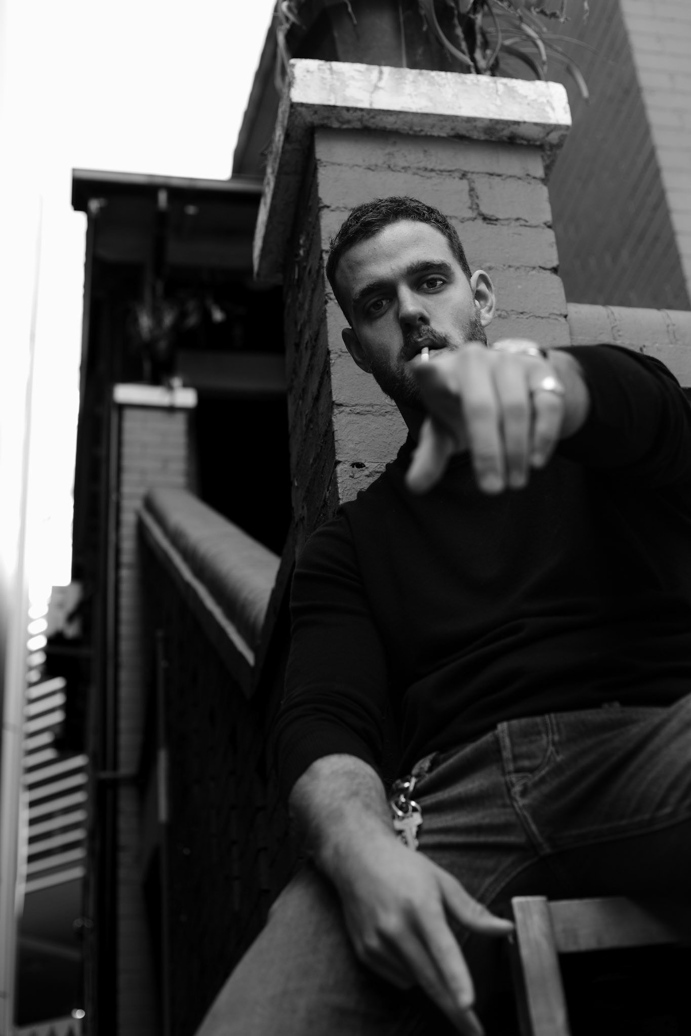 A black-and-white photo of a young man with short hair and a beard, leaning out from behind a brick wall with a serious expression, pointing towards the camera, wearing a dark shirt and jeans, with a keychain hanging from his pocket, and a cigarette 