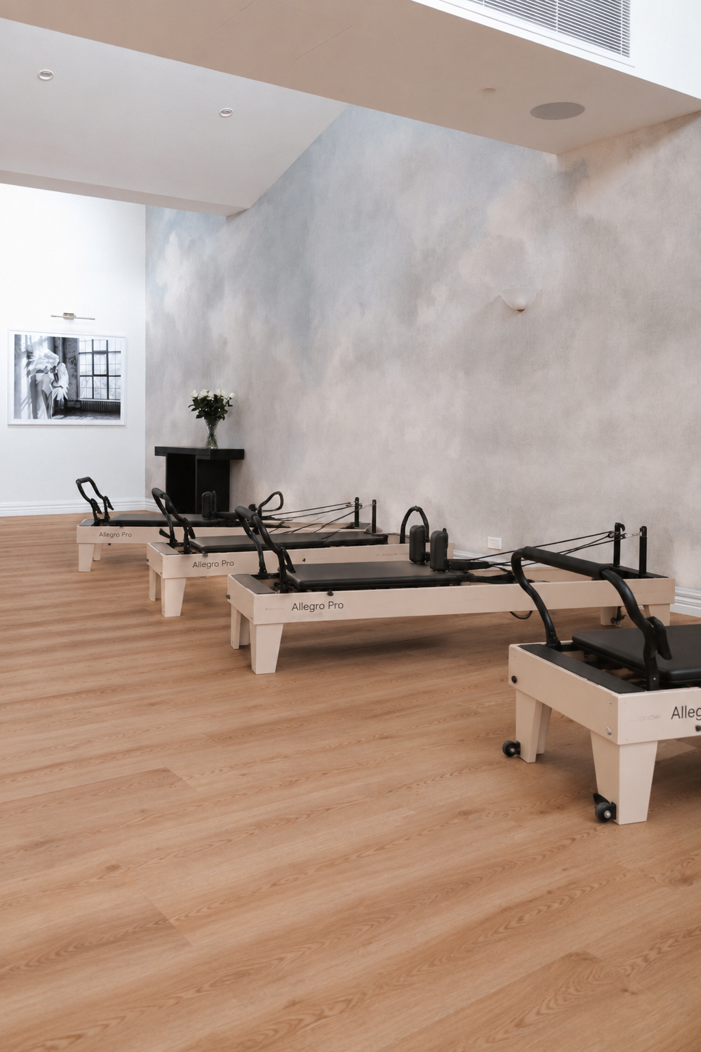 Four White Pilates reformer machines labeled 'Allegro Pro' arranged in a room with wooden flooring, a textured gray wall, a black fireplace with a vase of white flowers, and a black-and-white portrait picture on the wall.