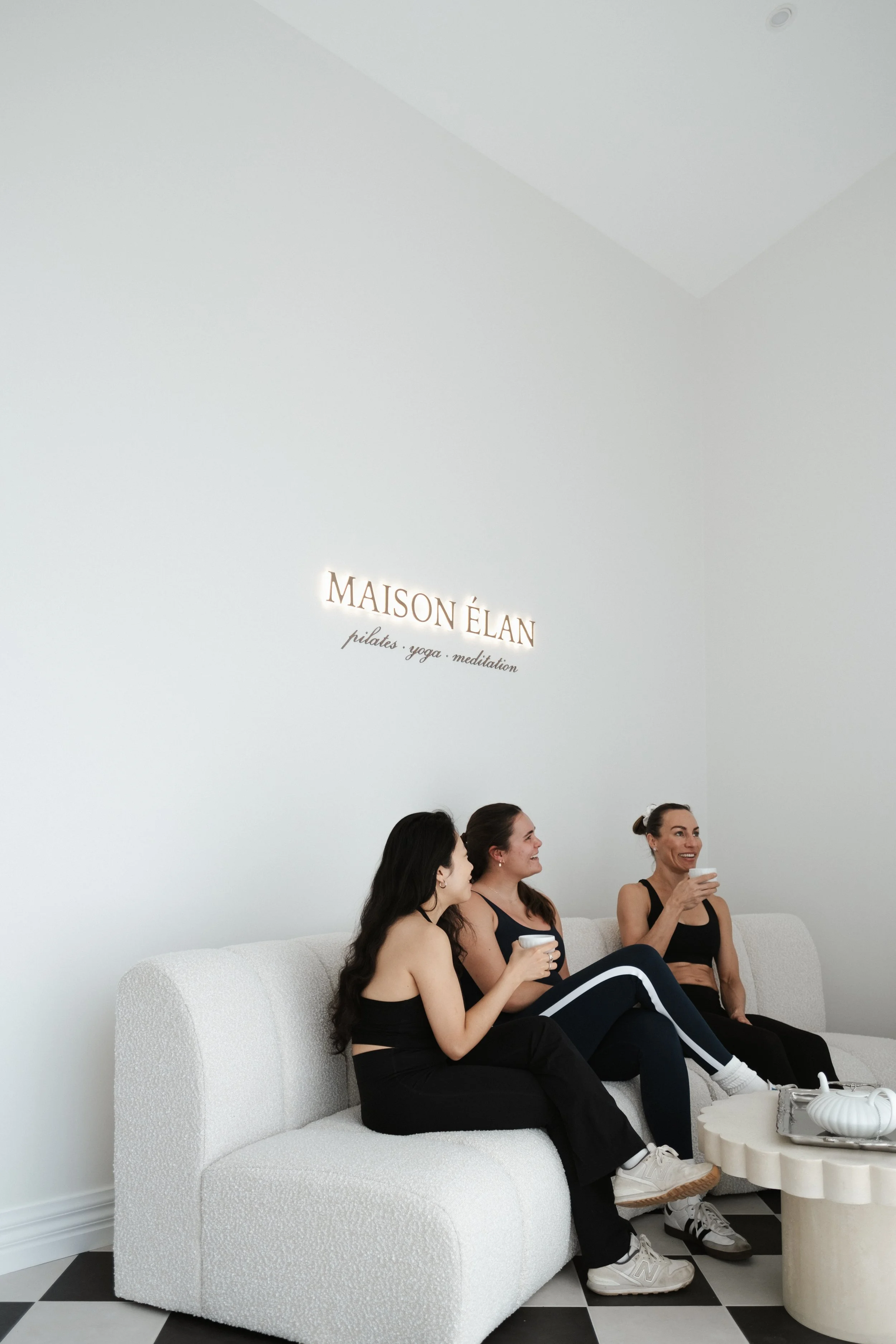 Four women sitting on a white couch, smiling and holding cups, in a minimalist room with a sign on the wall that reads 'MAISON ÉLAN pilates, yoga, meditation.'
