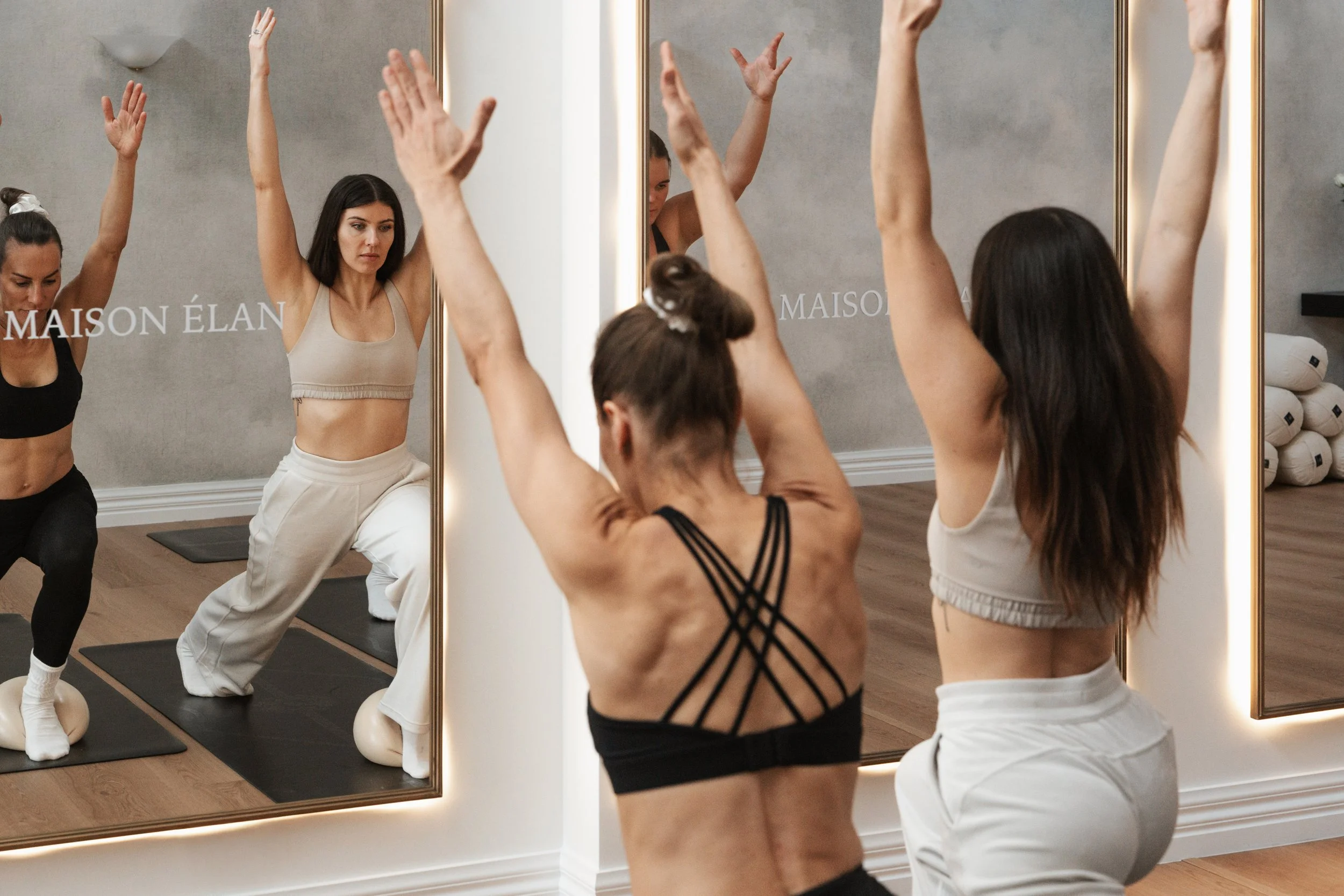 A group of women practicing yoga or dance in front of mirrors in a studio, wearing workout clothes, with their arms raised.