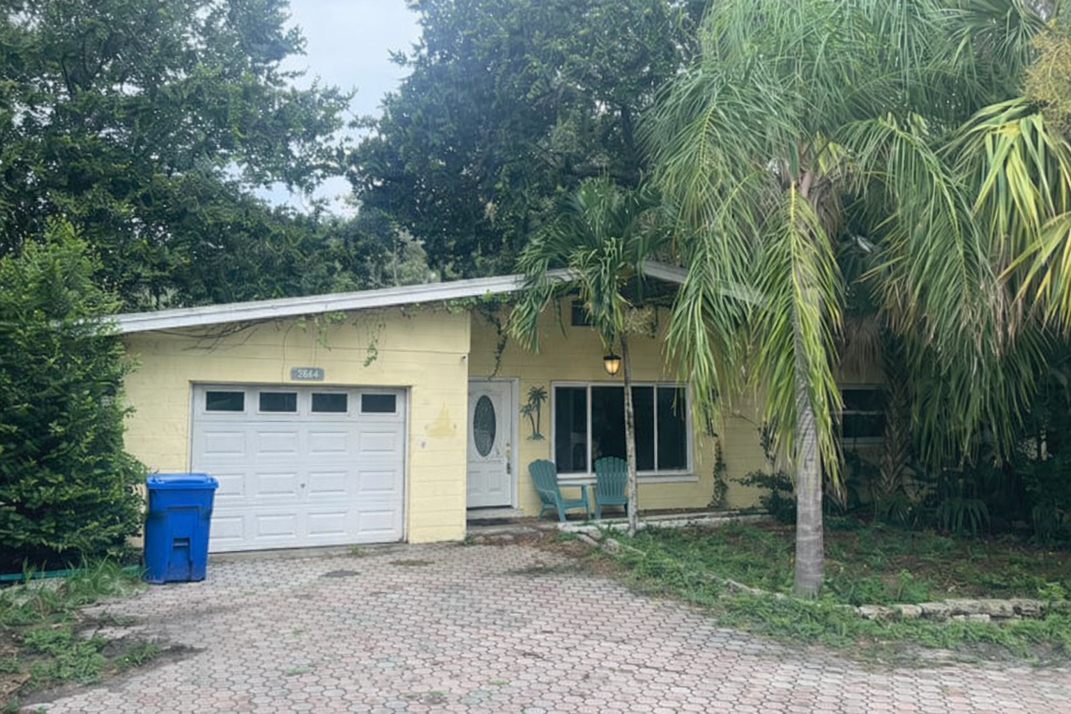 A yellow house with a white garage door, blue trash bin, and two teal chairs outside. The house has a large window and is surrounded by green trees and plants, including a tall palm tree.