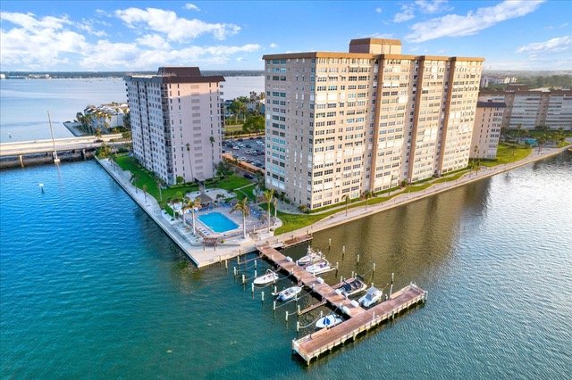 An aerial view of multi-story residential buildings next to a marina with several boats docked, water, and a blue sky with some clouds.