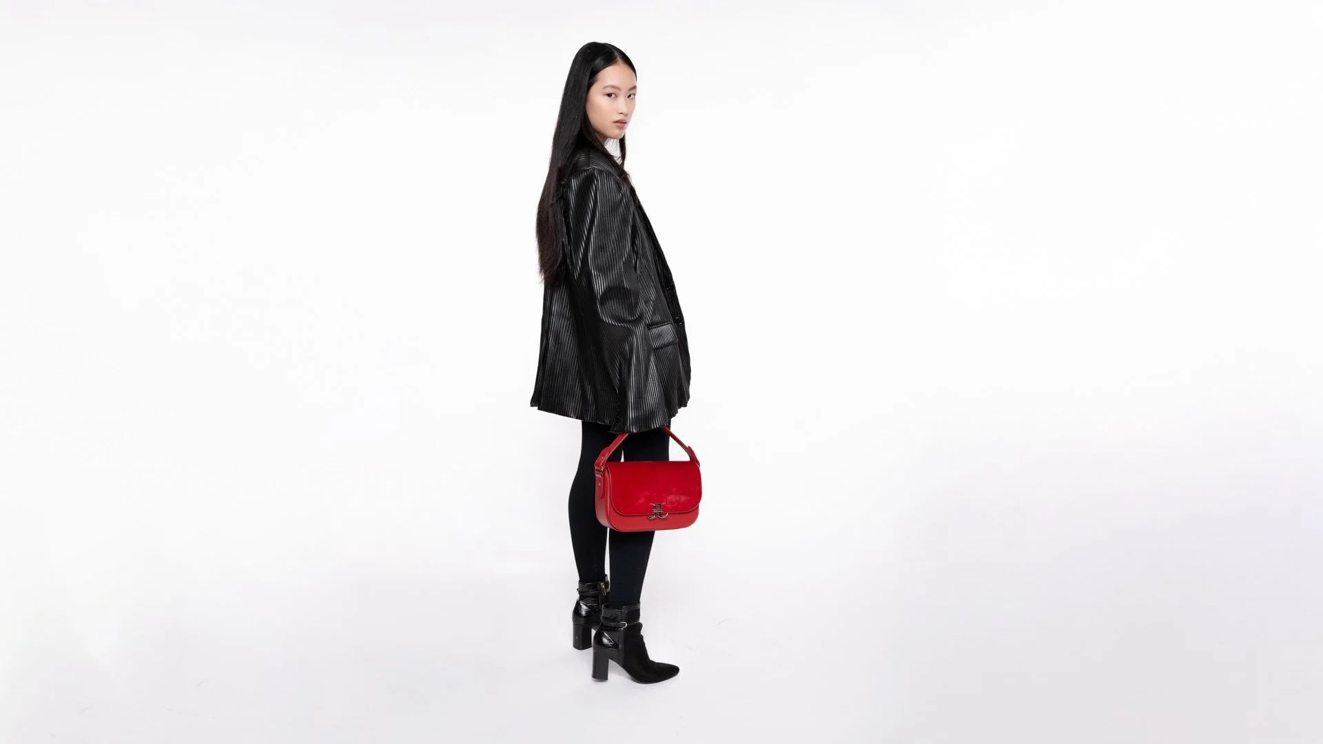 Woman with long black hair wearing a black leather jacket, black leggings, and black high-heeled boots, holding a red handbag, standing against a plain white background.