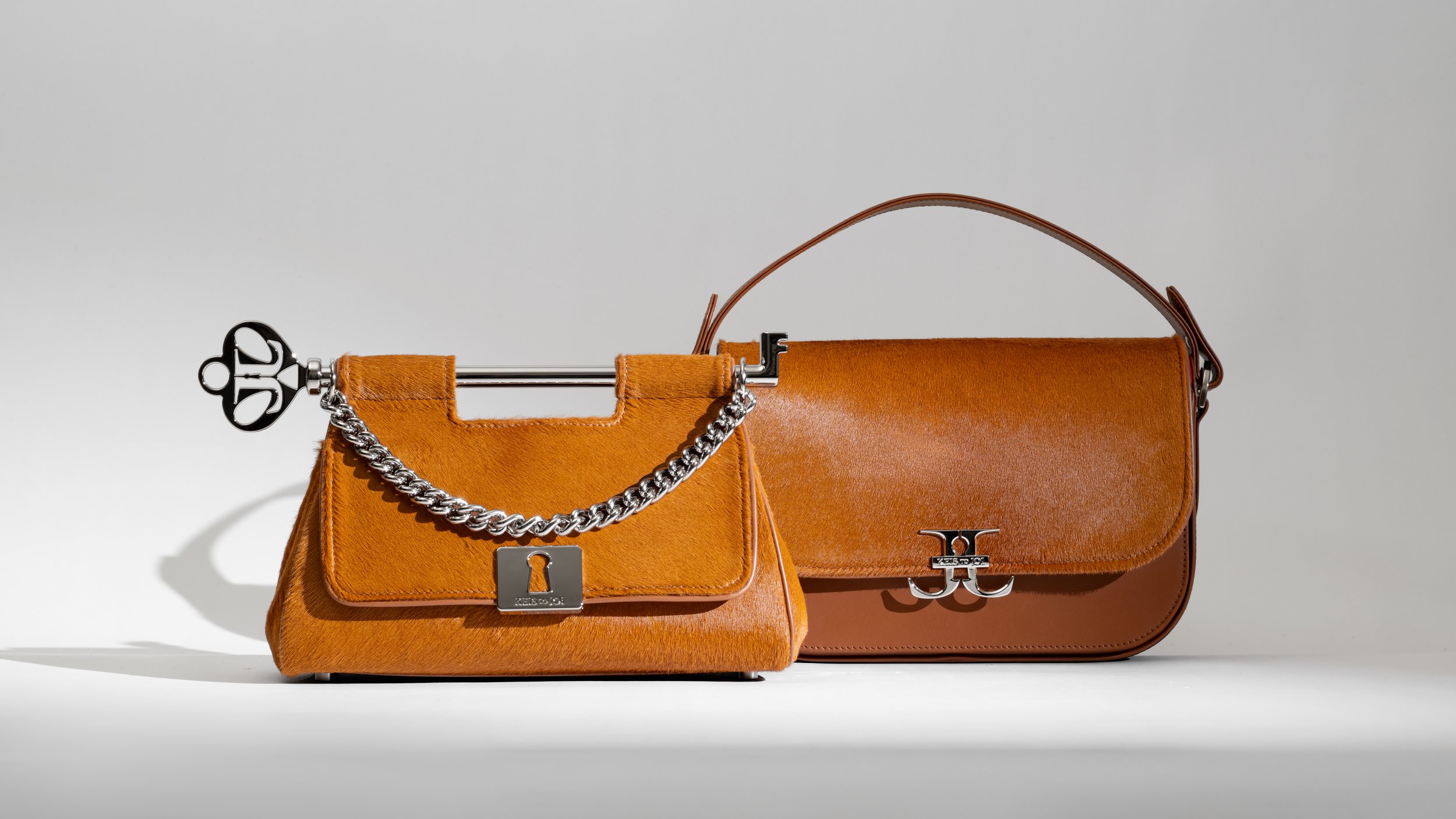 Two tan-colored handbags made of leather with silver hardware are displayed against a light gray background. The handbag on the left has a chain handle and a lock-shaped clasp, while the bag on the right has a rounded top handle and a logo clasp.