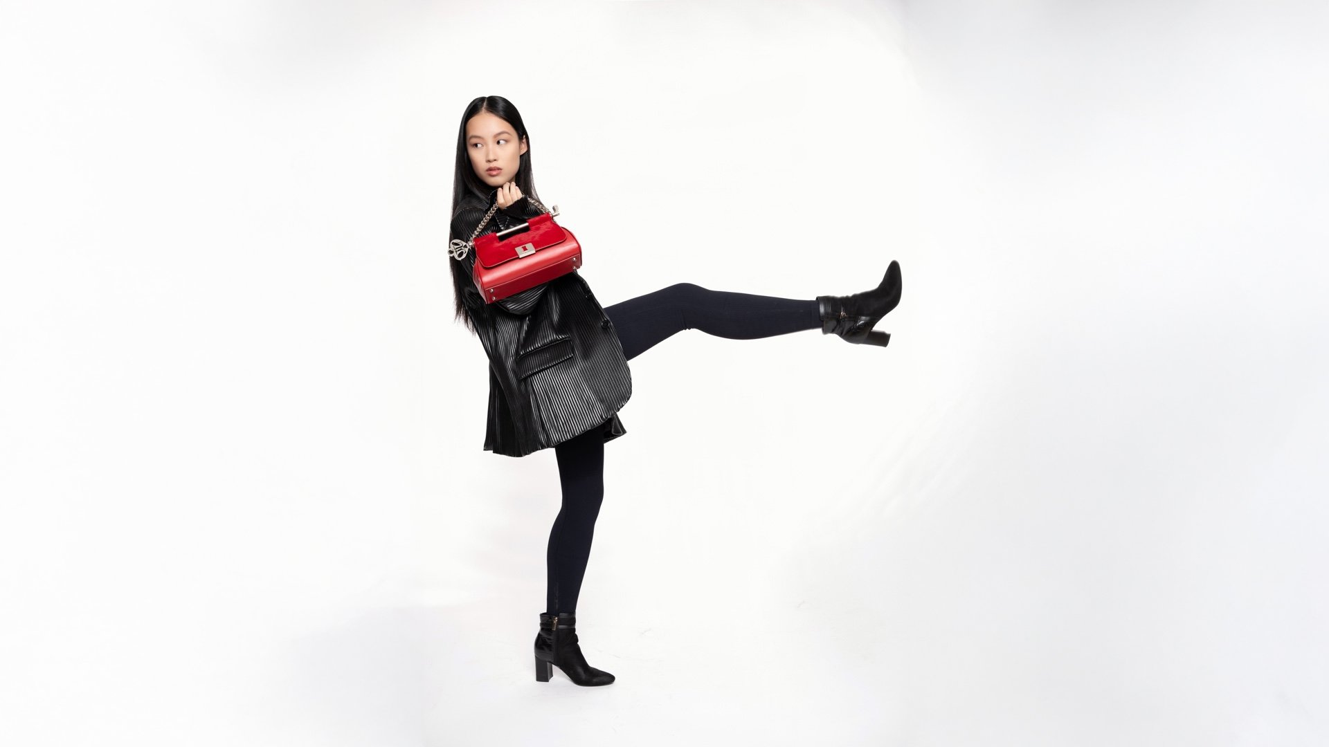 Young woman in black leather jacket, black leggings, black ankle boots, holding a red purse, raising one leg in a studio with a white background.