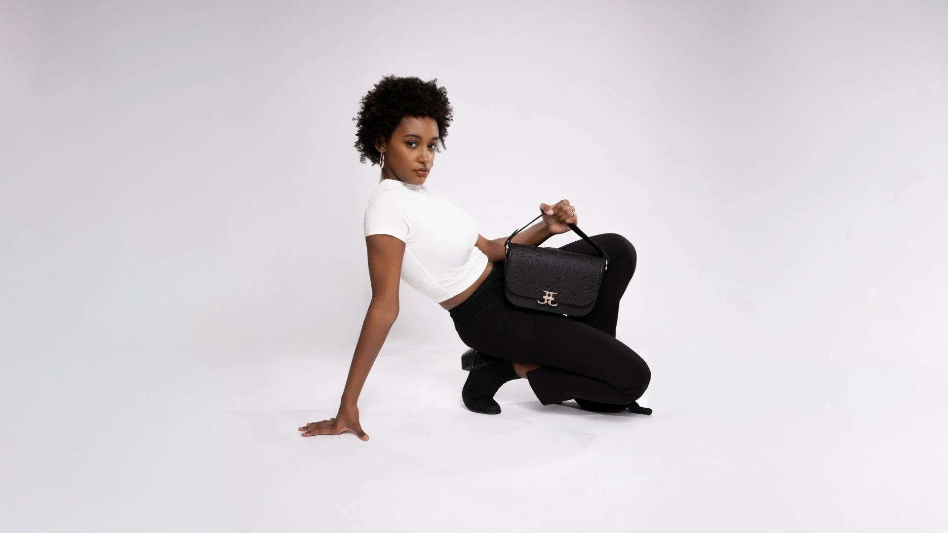A woman with curly hair kneeling on the floor, wearing a white crop top, black pants, and black shoes, holding a black purse with a gold logo.