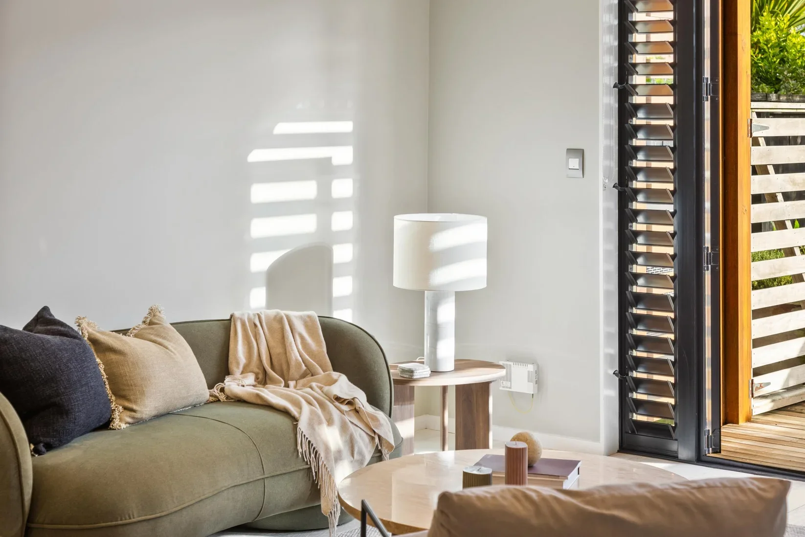 Living room with a green sofa, beige and dark pillows, beige throw blanket, white lamp on wooden side table, and sliding glass door with wooden slats leading to outdoor space.