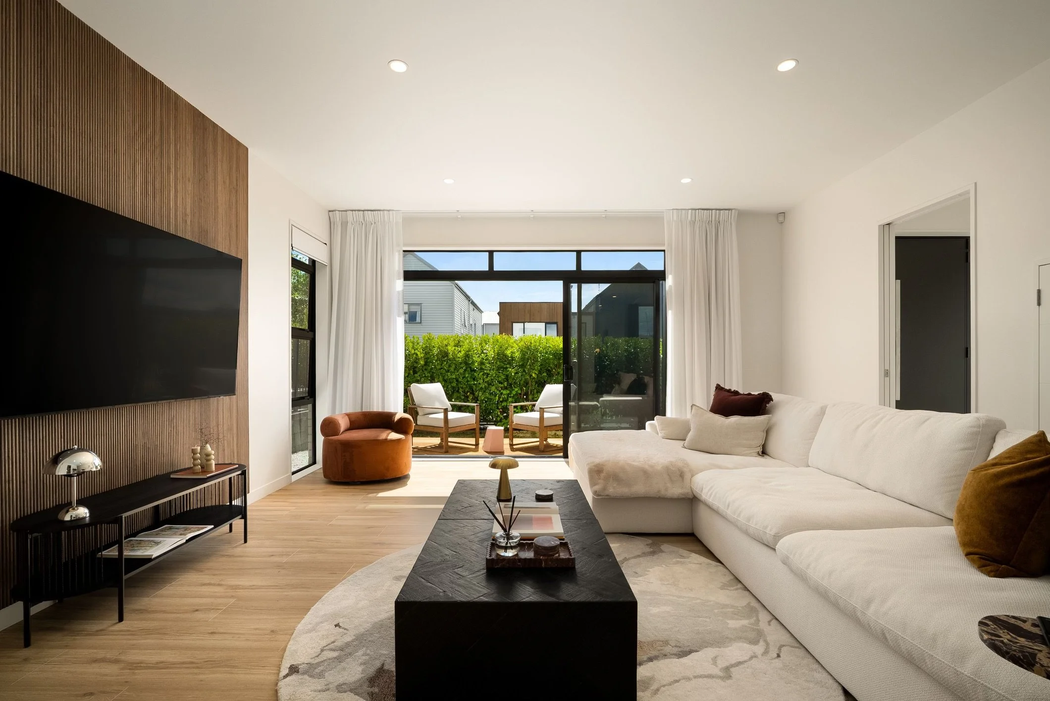 Modern living room with white sectional sofa, black coffee table, wall-mounted TV, and sliding glass door leading to an outdoor patio with two chairs and greenery.