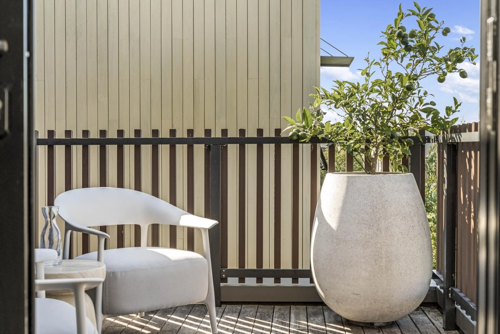 Small balcony with a white chair, a small table, a drinking glass, and a large potted citrus tree in a modern city setting.