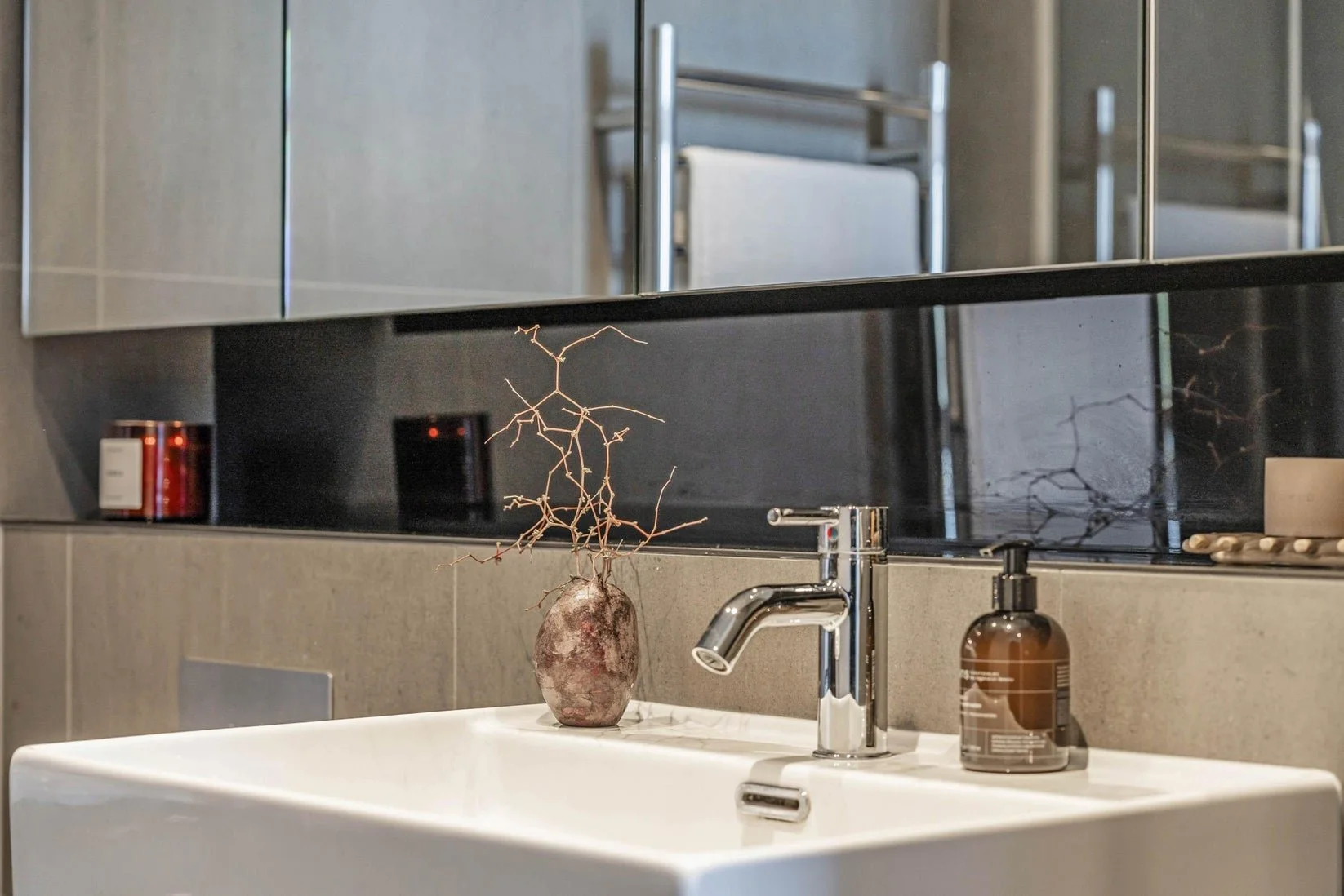 Bathroom sink with a vase holding a dried plant, soap dispenser, a mirror, and bathroom accessories on the countertop.