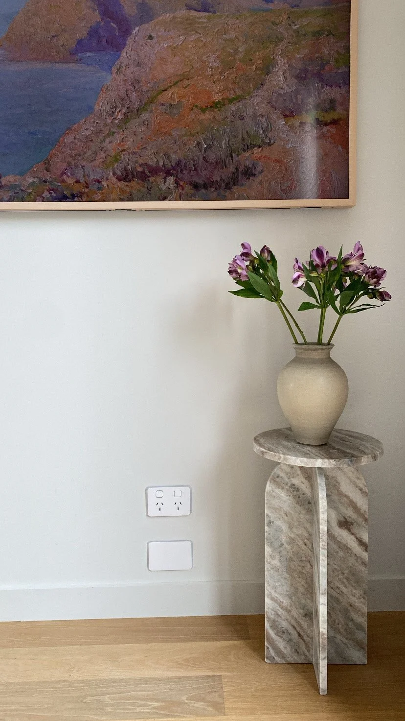 A beige ceramic vase with purple and white flowers on a marble pedestal table in a room with a painting on the wall and electrical outlets below.