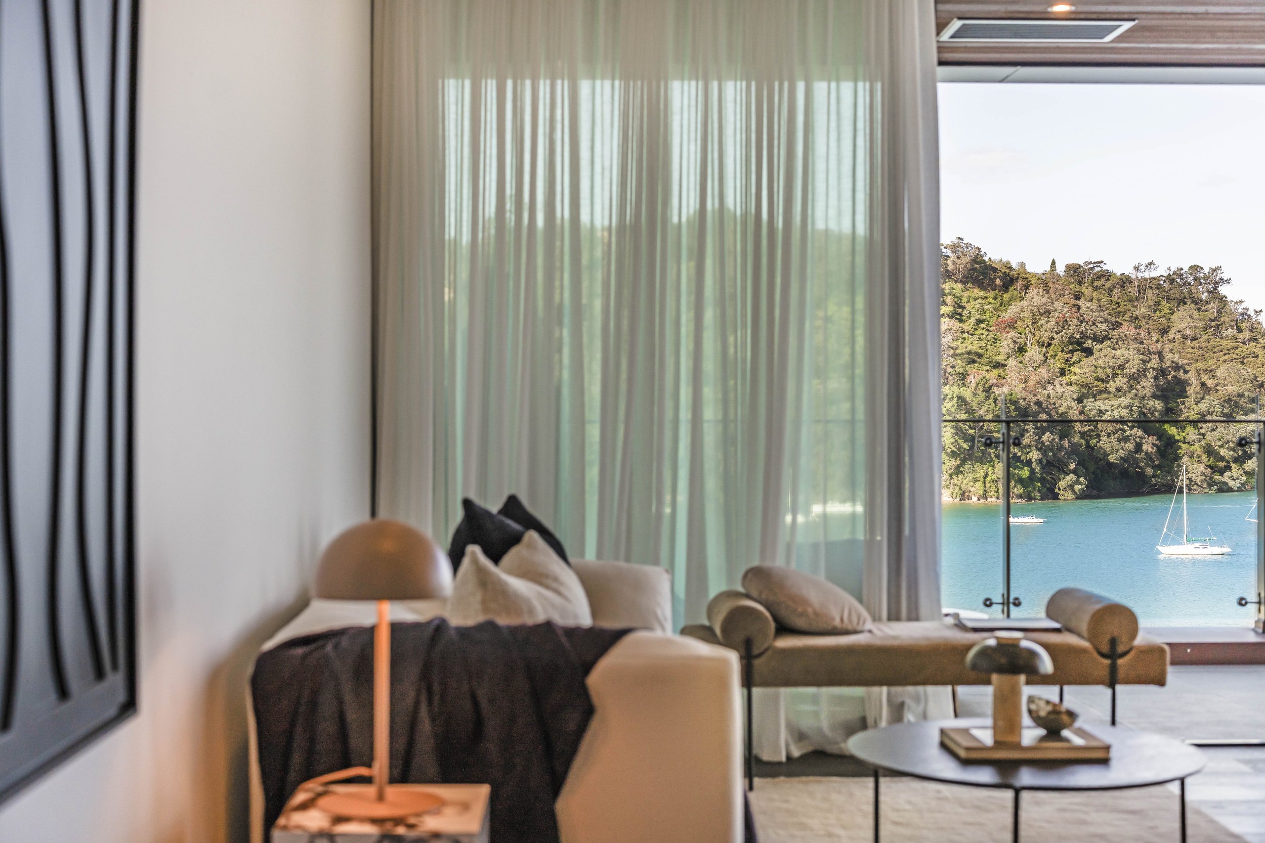 A cozy hotel room with a view of a lake and sailboats, featuring a beige sofa, pillows, a round marble coffee table, a modern table lamp, and sheer curtains.