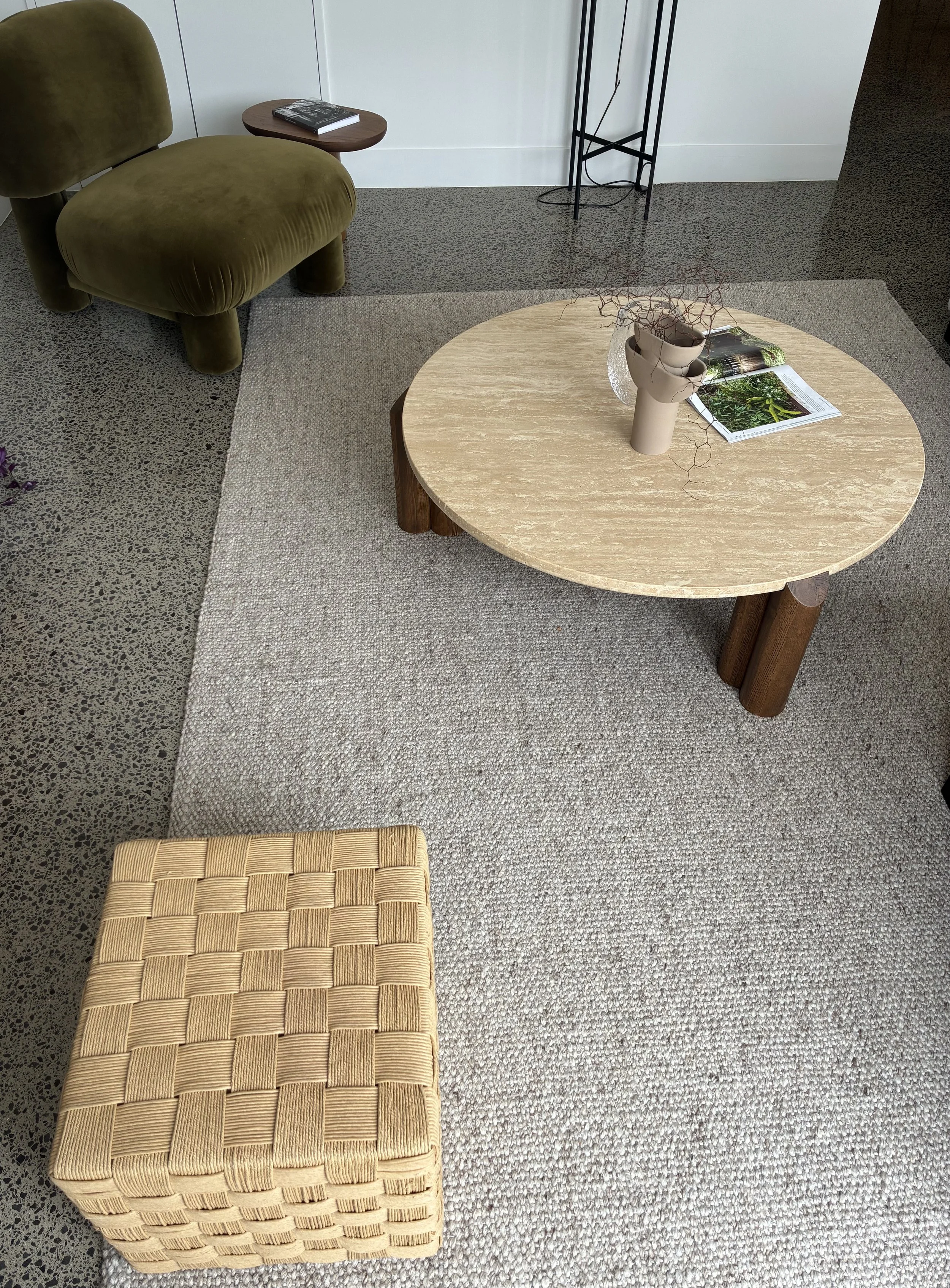 A round wooden coffee table with a beige vase containing dried branches and an open magazine, a mustard-colored woven square stool nearby, an olive-green velvet chair with rounded back and legs, a small wooden side table with a book, and a black plant stand, all on a beige woven rug in a room with white walls and a terrazzo floor.