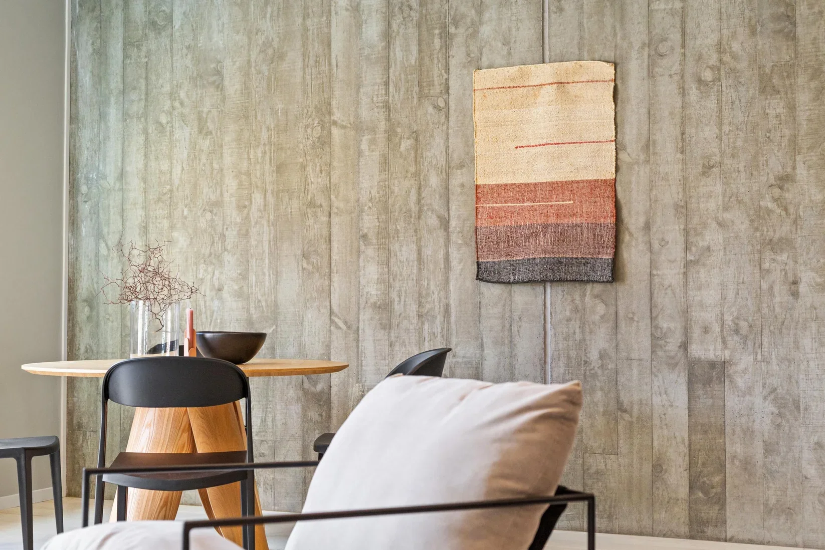 Modern dining space with a concrete wall, a small wooden table, black chairs, a beige pillow in the foreground, minimalist decor including a glass vase with a dried branch, a black bowl, and a textile wall hanging with horizontal stripes in beige, red, and black.