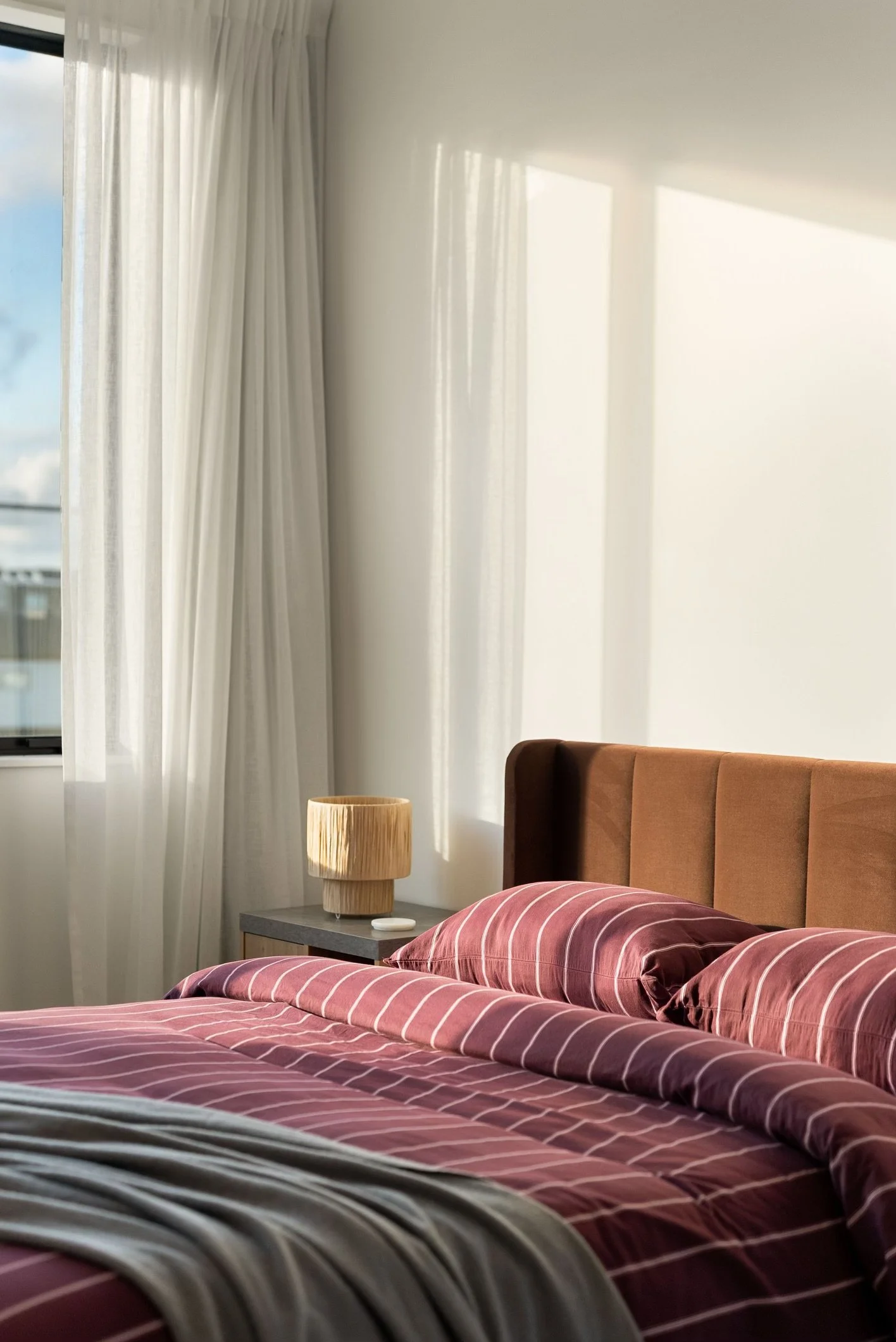 Sunlit bedroom with a bed that has a pink striped duvet and matching pillows, a tan upholstered headboard, a bedside table with a wooden textured lamp, and white curtains near a window.