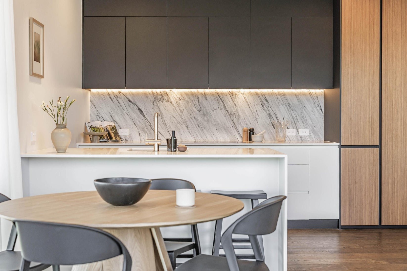 Modern kitchen with black upper cabinets, marble backsplash, white countertops, and a wooden dining table with black chairs.