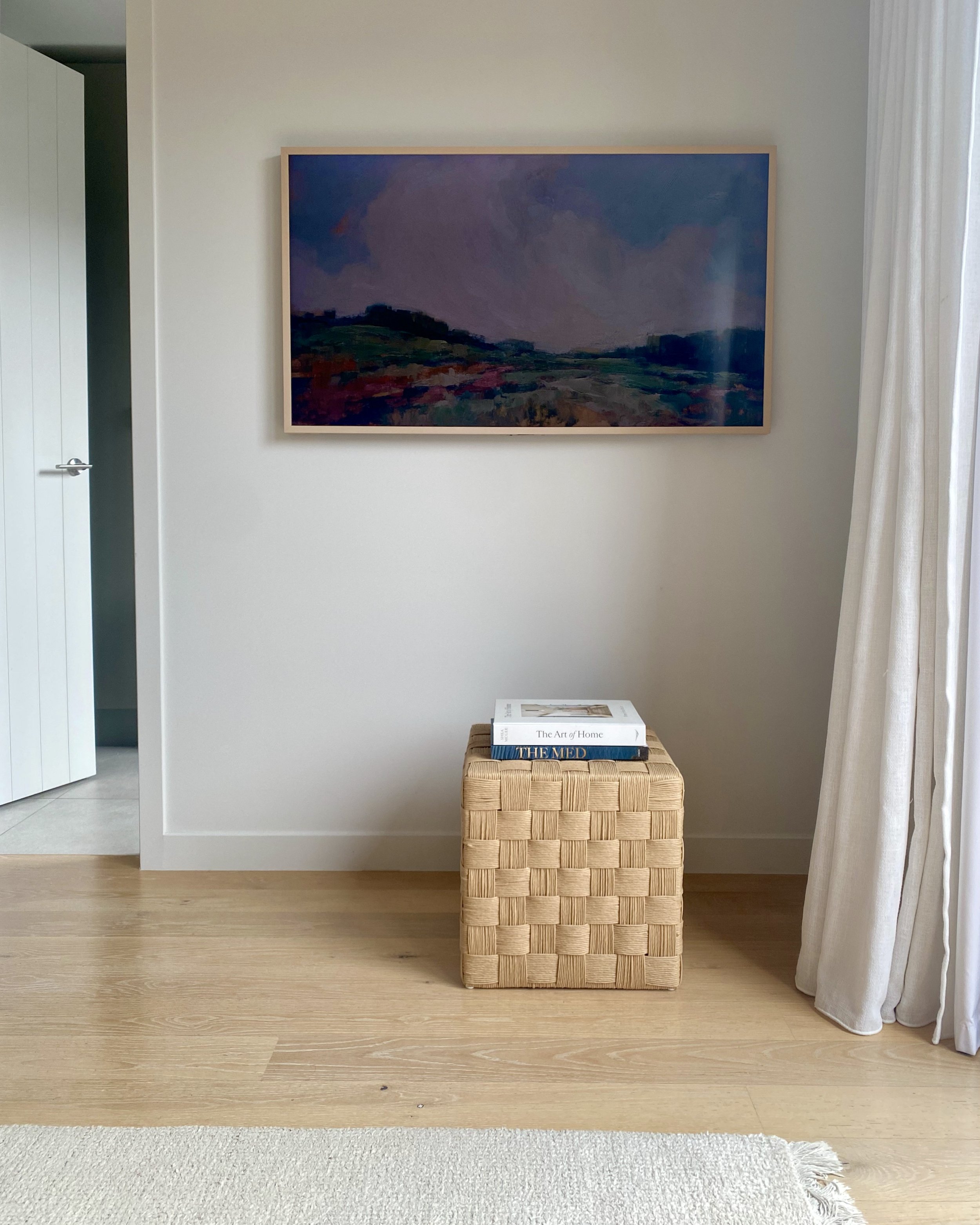 A room with a framed landscape painting on the wall, a woven wooden cube with three books stacked on top, a white curtain on the right, and part of a white door on the left.