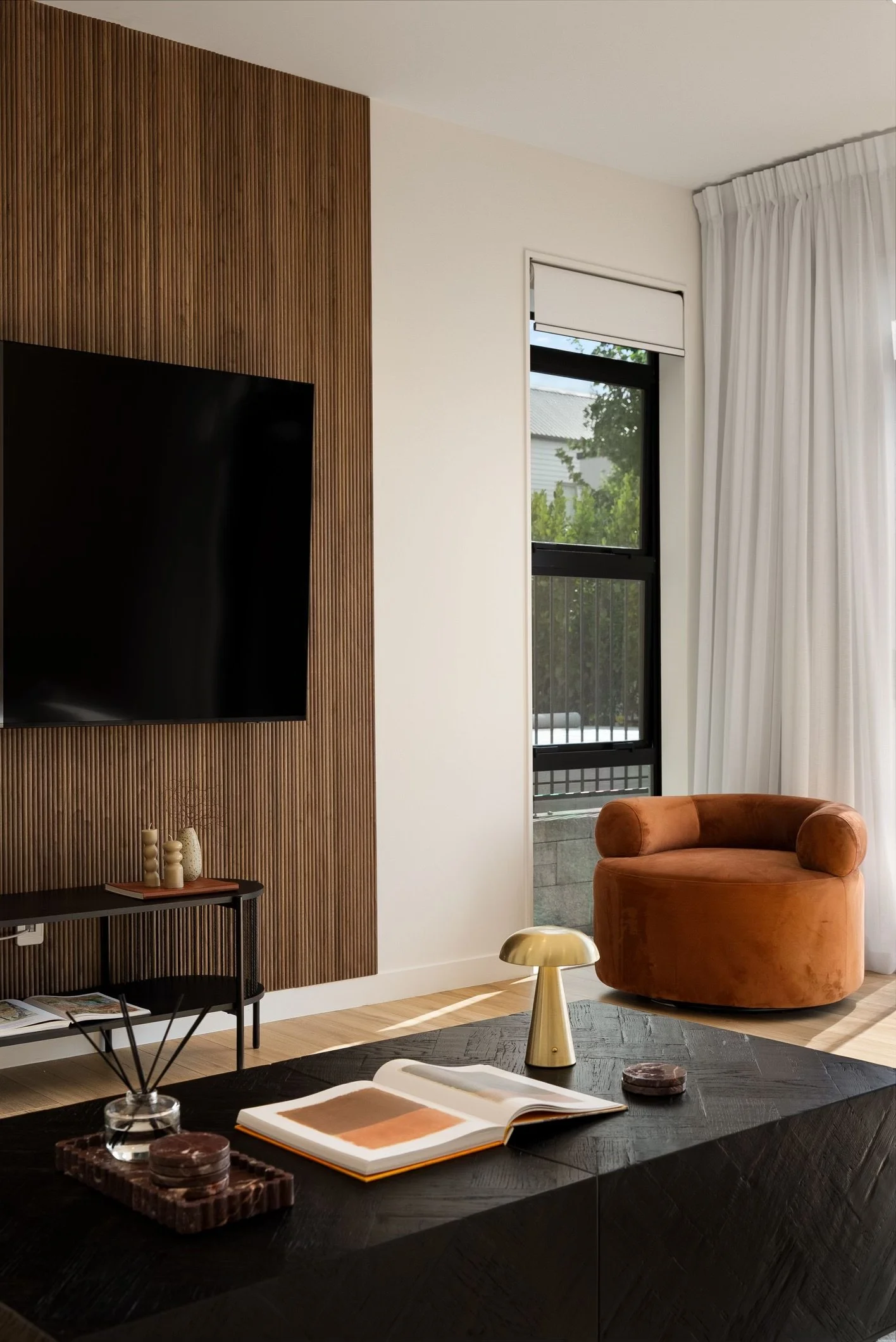 Modern living room with a large flat screen TV mounted on a textured wooden wall, a small black console table with decorative vases, a round brown armchair near a window with white curtains, a black coffee table with an open book, a small golden table lamp, and natural light streaming in through the window.