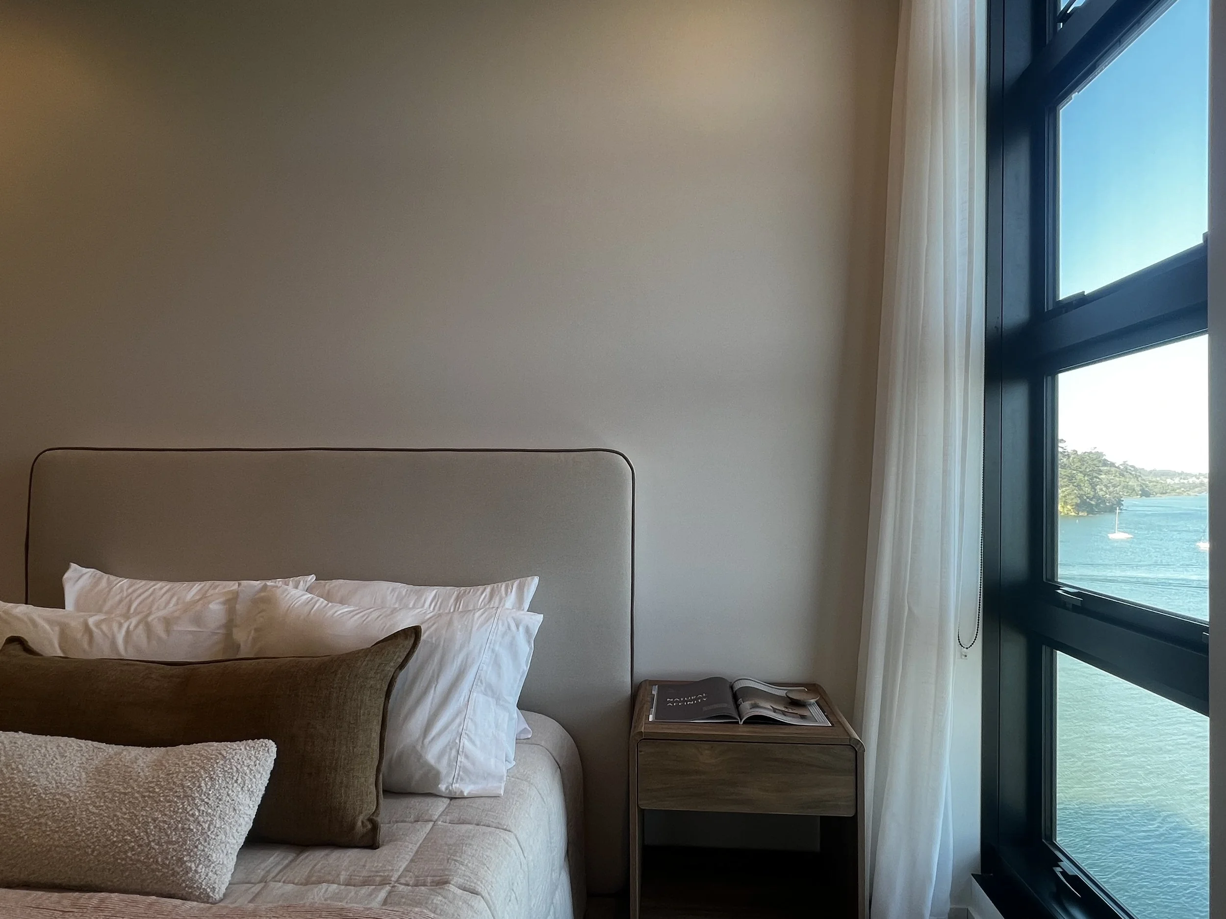 A bedroom with a bed, pillows, and a bedside table next to a window overlooking a lake with trees and boats.