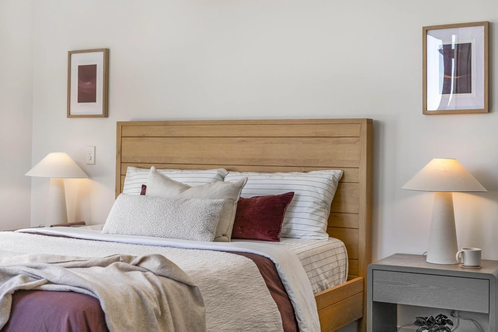 A bedroom with a wooden headboard, striped and solid pillows, a folded blanket, two table lamps on nightstands, and framed artwork on the white wall.