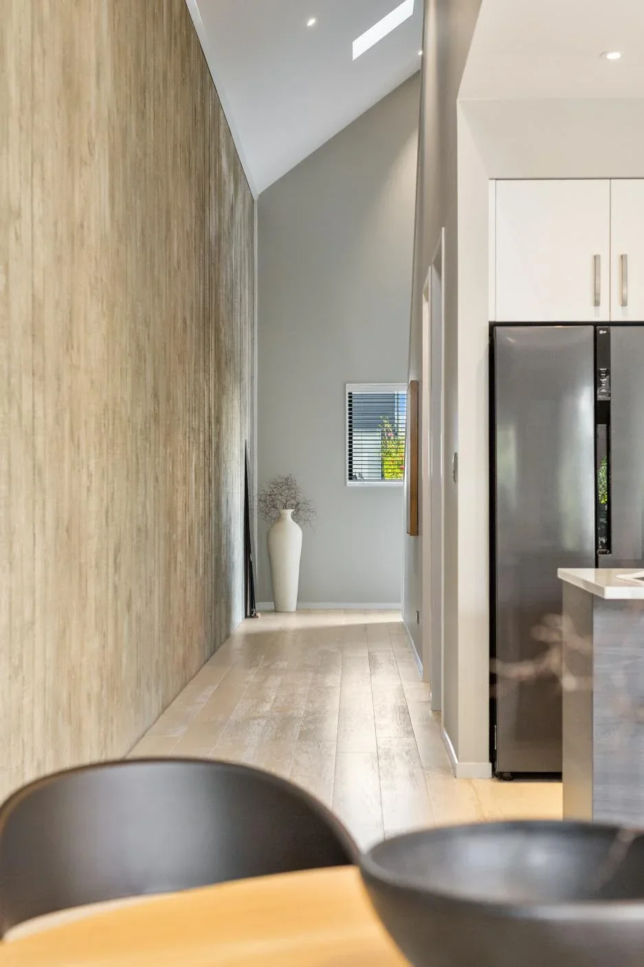 Interior view of a modern kitchen and hallway with light-colored wooden floors, white cabinets, stainless steel refrigerator, a large vase with decorative branches, small window with blinds, and part of a dining table with black bowls in the foreground.
